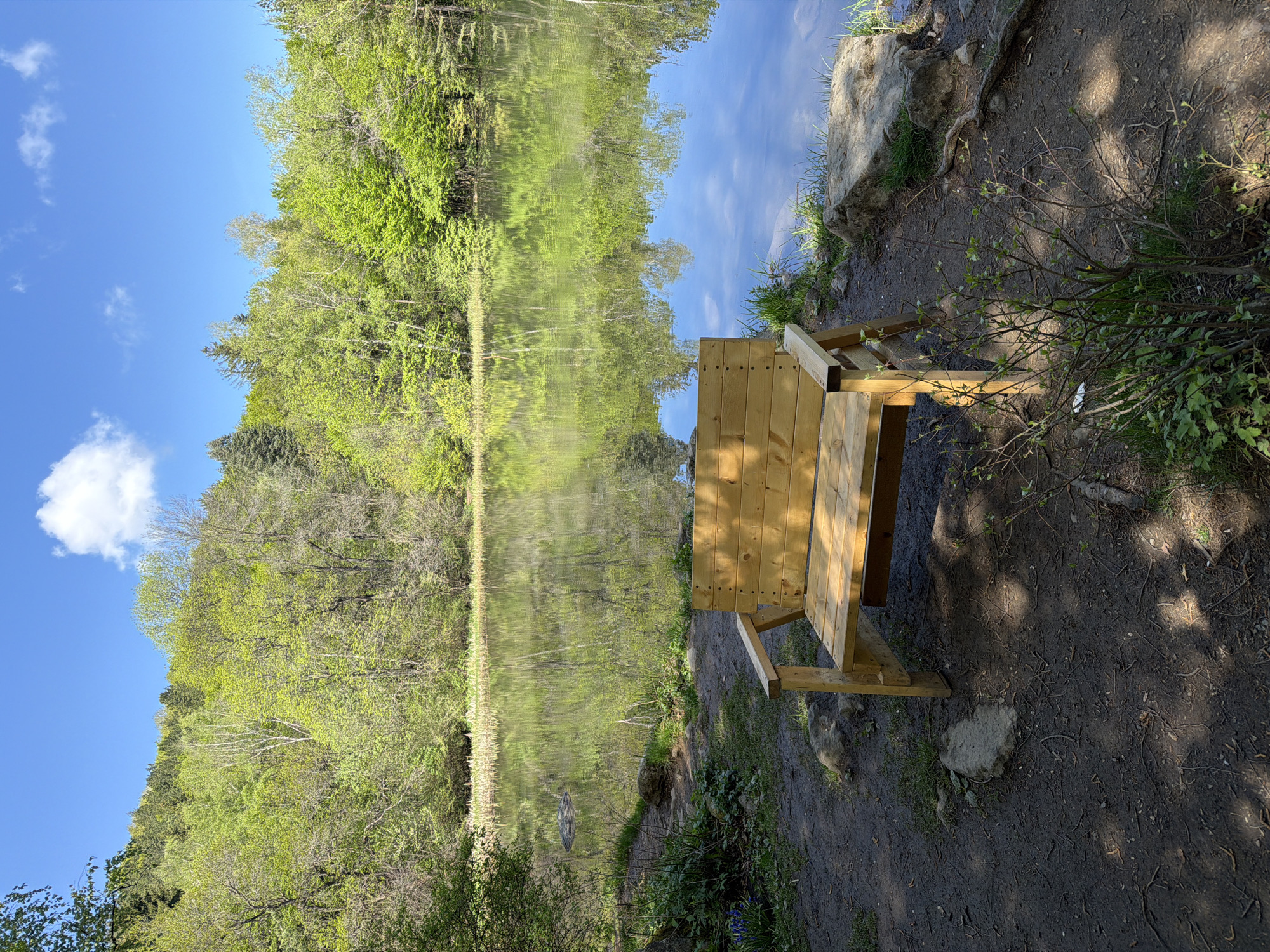 A wooden bench is located on a dirt path near a body of water, likely a lake or pond, in Sofia, Bulgaria. Trees with green foliage line the opposite side of the water, creating a reflection in the water's surface. The sky is visible above the trees, with scattered clouds. The ground around the bench is covered in dirt and some vegetation.