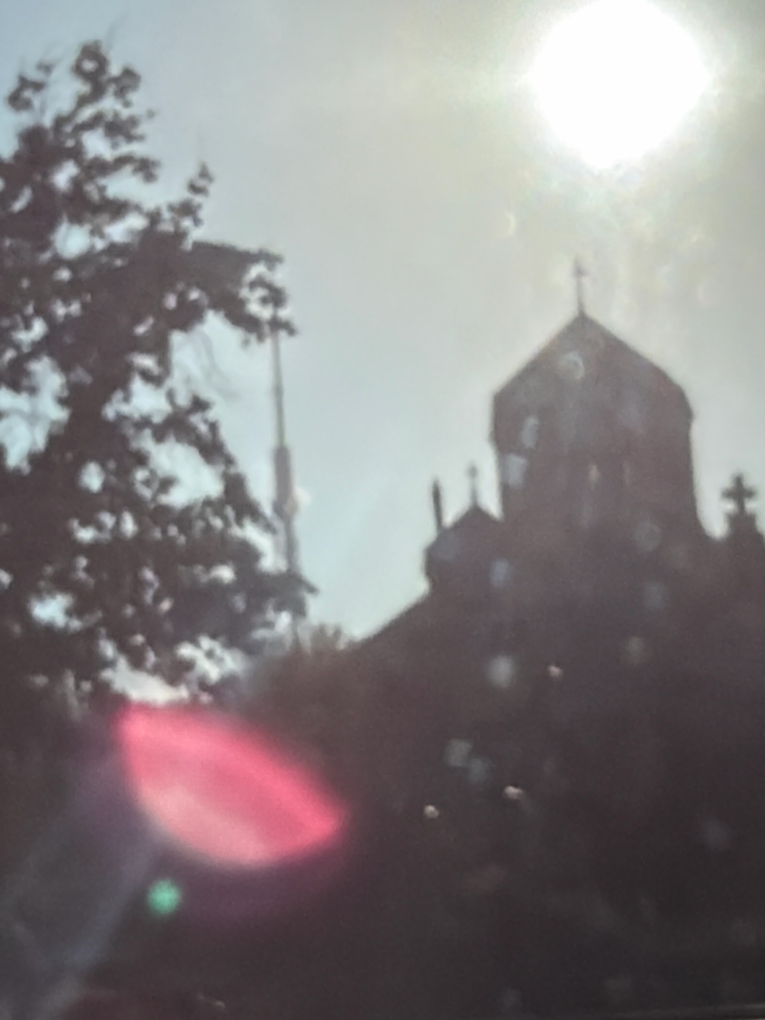 The image captures a silhouetted view of a church, likely St. Gregory the Illuminator Cathedral, in Yerevan, Armenia. The sun is bright and high in the sky, with visible lens flare creating a halo effect. In the foreground, to the left, is the dark silhouette of a tree, its branches creating a textured canopy. Behind the tree and to the right of the church, a tall, slender structure, possibly a radio tower or antenna, rises against the hazy sky. The overall scene is characterized by low light conditions and a somewhat hazy atmosphere, with bright reflections and a prominent sun disk. There are no people or animals visible in the image.