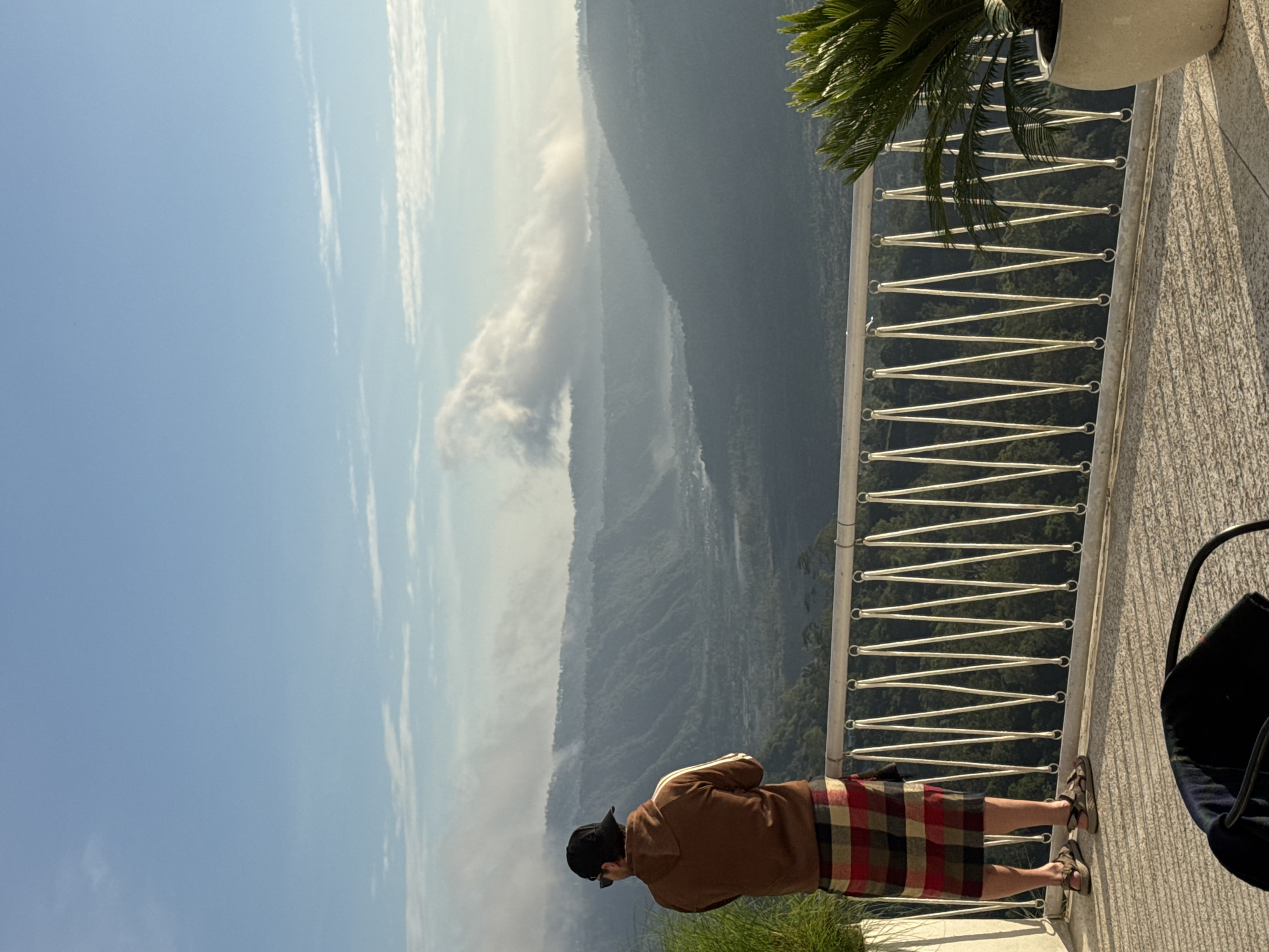 A full-body view captures an individual on a balcony overlooking a mountainous landscape. The person is wearing a brown jacket, a plaid skirt, sandals, and a black baseball cap. The balcony features a white railing, and plants in a white pot. In the background, a large mountain range extends under a hazy blue sky. The photo suggests the location might be in Kedisan, Indonesia, given the landscape features.