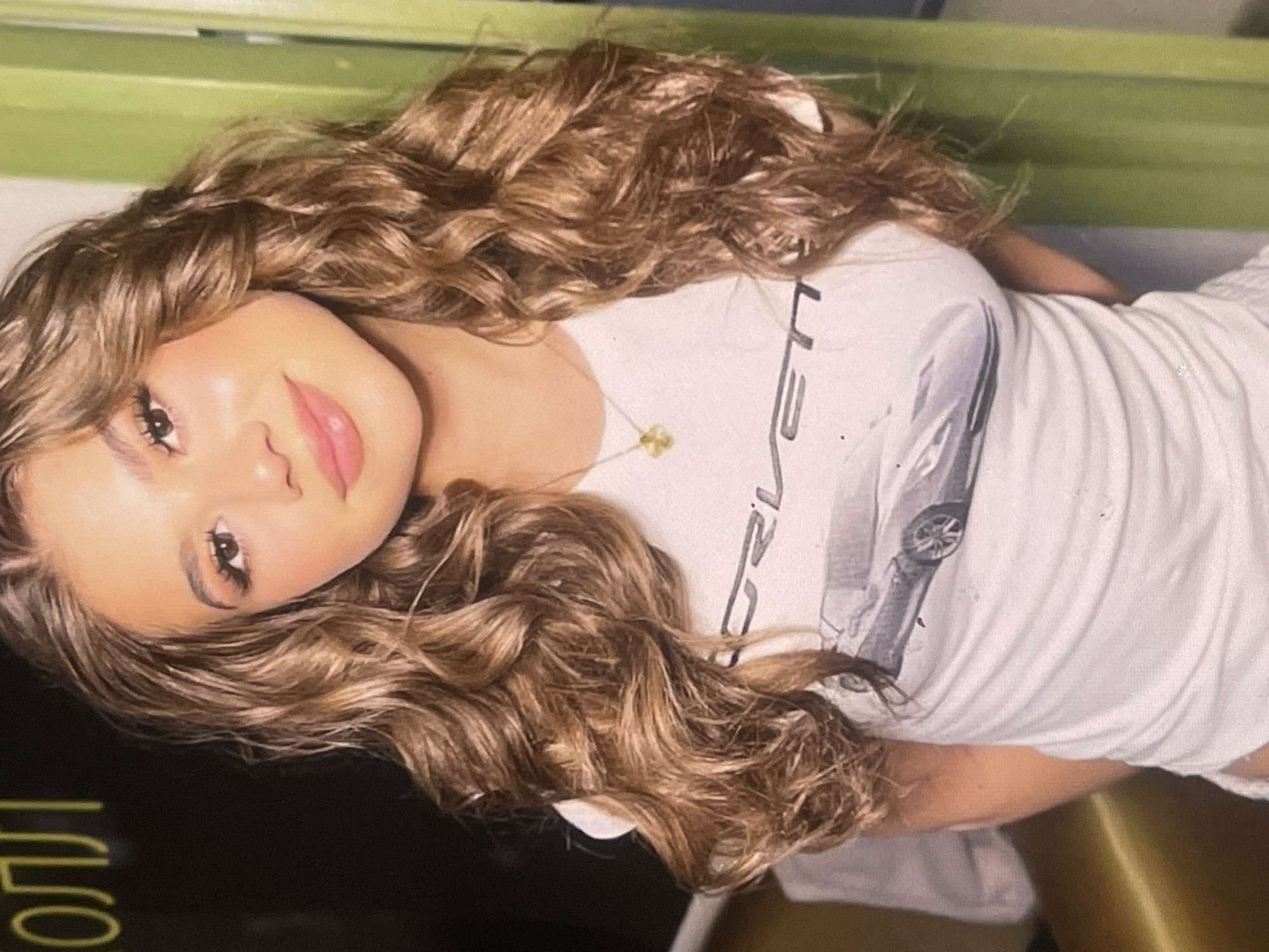 The image captures a young woman in a close-up, horizontally oriented shot, likely taken indoors in Poltava, Ukraine. She has long, voluminous, curly light brown hair that cascades around her. Her face is turned towards the viewer, featuring prominent eye makeup with dark lashes and pink lipstick on her full lips, accompanied by a gentle smile. She is wearing a white short-sleeved t-shirt with a graphic print on the front. The print prominently features the partial word "CORVETTE" in black text, along with a grey-toned image of what appears to be a Corvette C8 sports car. A delicate gold-colored necklace with a small, intricate pendant (possibly a flower shape) is visible around her neck.
The background consists of two main elements: a light green surface or wall in the upper portion and a dark, almost black, surface on the lower left. On this dark surface, glowing yellow-green Cyrillic text is partially visible, spelling out "ПОЛЛ" (POLL), which could be a sign or part of the décor, possibly hinting at a specific venue in Poltava. The woman appears to be posing for the camera, conveying a calm and pleasant demeanor. The lighting suggests an indoor setting, and no specific time of day or weather conditions are discernible from the image itself.