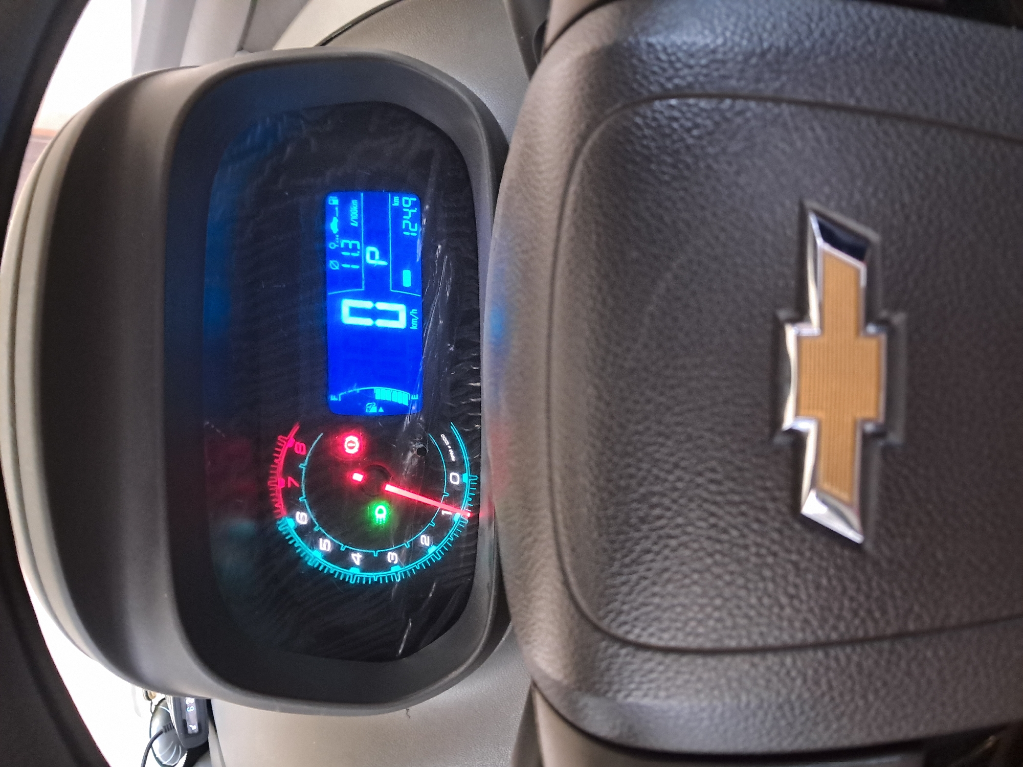 The image shows a close-up of a car's dashboard and steering wheel, with a Chevrolet logo visible on the steering wheel. The dashboard displays a digital speedometer showing "0 km/h", fuel gauge, and trip odometer reading "124.9 km". There is also a tachometer with a red needle pointing to "0". The car is parked in a city, but the exact location in Bagish, Uzbekistan is not specified in the image.
