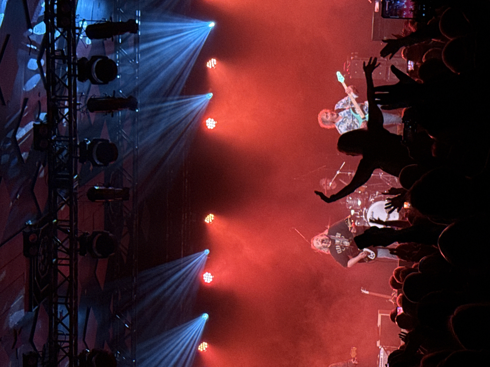 The image depicts a live music concert within an indoor venue in Unknown, Unknown. The scene focuses on an illuminated stage, viewed from the audience perspective. Overhead, a metal truss structure supports multiple stage lights emitting broad red illumination and distinct blue light beams that cut through atmospheric haze.
On stage, two male musicians are prominently visible. To the left, a man with long, light-colored hair, wearing a dark t-shirt, is playing an electric guitar and singing into a microphone. To the right, another man with shorter, curly light-colored hair, dressed in a patterned shirt, plays a light blue electric bass guitar. A drum kit is partially visible behind the first guitarist, and an additional guitarist is partially obscured to the far left of the stage. Stage equipment visible includes guitar amplifiers and microphone stands.
The foreground is filled with a dense crowd of audience members, mostly silhouetted against the bright stage lighting. Many individuals have their hands raised in the air. One prominent silhouetted figure in the mid-ground has both arms extended upwards. At least two audience members are holding up smartphones, one clearly displaying its screen as it records the performance.