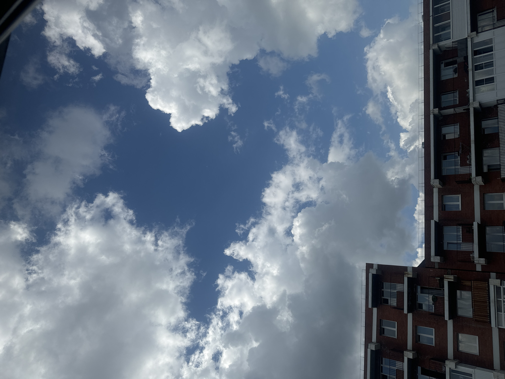 The image depicts the facade of a brick apartment building against a bright blue sky filled with white and gray clouds. The building has multiple stories, each with numerous windows and balconies with black railings. The architecture appears modern, with clean lines and white trim around the windows and balconies. The sky is dynamic, with large, fluffy cumulus clouds interspersed with thinner, wispy cirrus clouds. The overall impression is of a sunny day with variable cloud cover. This scene is characteristic of residential areas in cities like Маркова, Russia, where such apartment buildings are common. There are no people visible in the image.