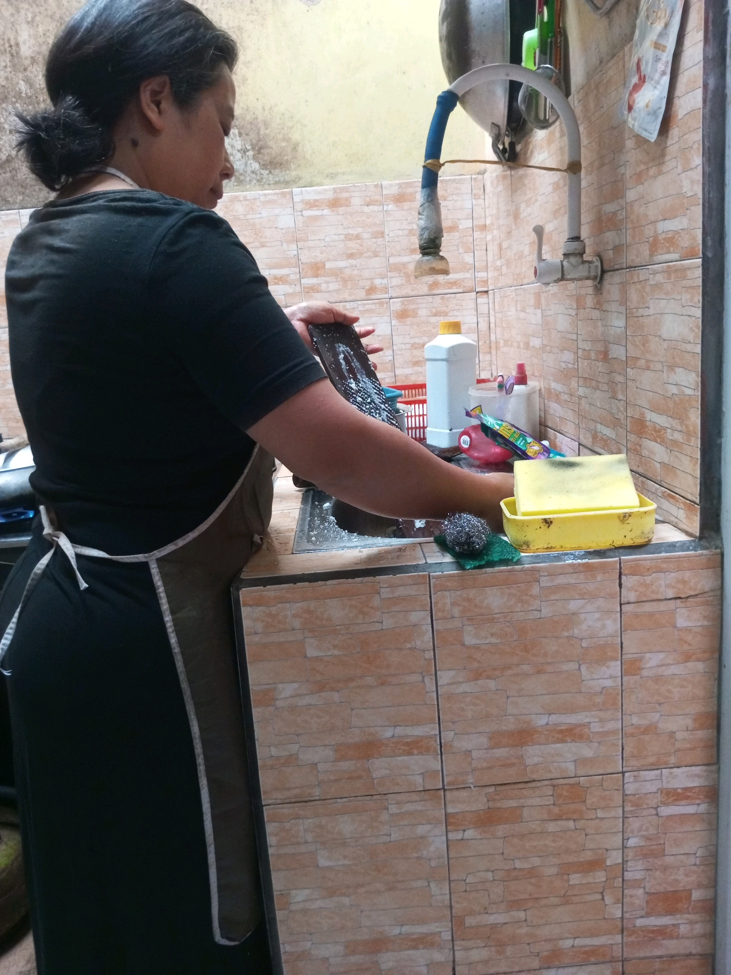 A woman is washing dishes at a sink. She is wearing a black t-shirt and an apron. The sink is made of stainless steel and is filled with soapy water. There are several bottles of dish soap and a yellow sponge next to the sink. The walls are tiled with a light brown brick pattern. The faucet is white and has a blue hose attached to it. The overall setting appears to be a domestic kitchen.