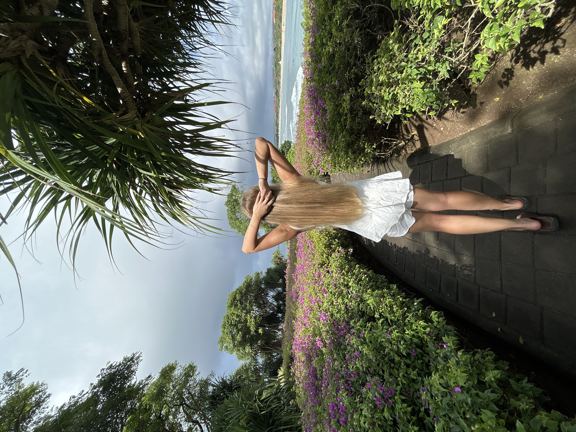 The image displays a full-body view of a person from behind, standing on a paved pathway and gazing out at a tropical coastal scene. The subject is a woman with very long, straight blonde hair, wearing a white sleeveless dress and dark flip-flops. Her arms are raised with her hands clasped behind her head.
The scene is set outdoors amidst lush tropical vegetation. To the right of the pathway, a dense hedge of green bushes bursts with numerous small purple flowers, possibly bougainvillea, extending towards the horizon. To the left, large green, spiky palm-like leaves frame the foreground from an overhead tree. Beyond the immediate greenery, a wide vista of the ocean unfolds, featuring light blue water with white foamy waves breaking on a sandy beach. A distant coastline with faint structures is visible on the horizon.
The sky above the ocean is mostly overcast with patches of brighter light, suggesting daytime, possibly a cloudy day or late afternoon. The pathway is made of dark, interlocking pavers, creating a neat border alongside the flowering bushes. No specific activity is taking place beyond the person standing and observing the view. There is no visible text in the image. The combination of tropical plants, a sandy beach, and ocean suggests a warm, coastal resort-like environment.