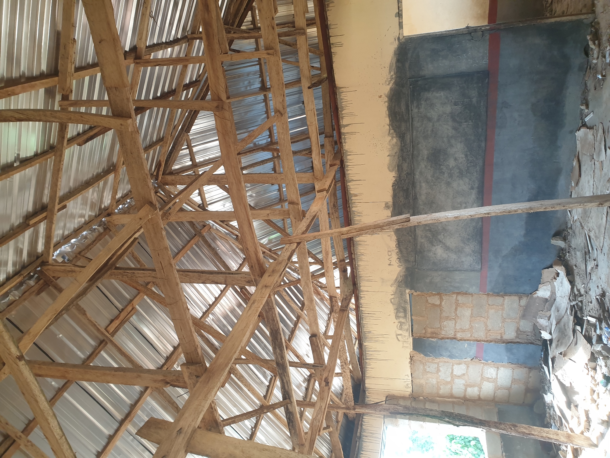 The scene, located in Aroro, Nigeria, is an interior space in a state of construction or disrepair. An exposed roof structure features a rudimentary wooden truss system supporting corrugated metal sheets, with visible gaps allowing ambient light to penetrate. The upper sections of the walls are painted a light beige and display numerous dark, vertical drip stains originating from the ceiling line. A horizontal red band separates these from the lower wall sections, which are painted dark blue-grey. Portions of the walls are unfinished, revealing bare concrete block construction, notably around a doorless frame. A distinct dark grey rectangular panel, possibly a chalkboard, is present on one wall. The concrete floor is covered with an accumulation of construction debris, including fragments of concrete, wood, and other materials, alongside scattered refuse. Multiple unprocessed wooden beams serve as visible structural supports, extending from the floor to the roof truss system. No individuals, actions, or interactions are observed within the frame.