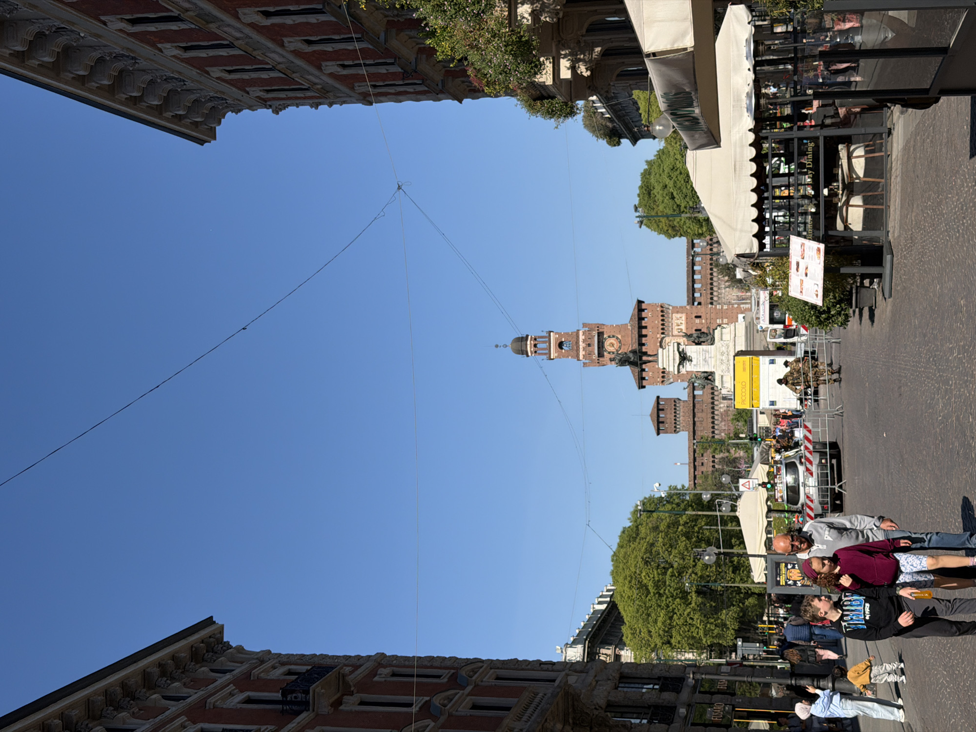 The image depicts a scene in Milan, Italy, with the Sforza Castle visible in the background. The sky is clear and blue. Power lines stretch across the upper portion of the image. Buildings flank both sides of the frame. On the right side, a pedestrian area features people walking and reclining. There is outdoor seating for a restaurant or cafe. In the distance, soldiers and a utility truck stand in the street next to the Castello Sforzesco, a medieval castle.