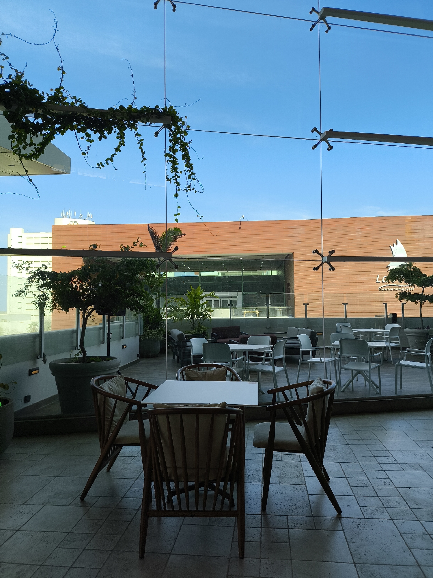 The image depicts a restaurant or cafe space, likely located on an elevated terrace or within a glass-walled structure in Porlamar, Venezuela. Inside, there are tables and chairs, some made of wood and others of plastic. Outside, a large brick-colored building is visible through the glass. The building has the word "La" written on it, part of a longer title. The sky is blue and visible through the glass and above the building. Green plants, including vines, decorate the indoor space. The floor is tiled with gray square tiles. No people are visible in the immediate foreground or background of the scene. The lighting suggests it is daytime.