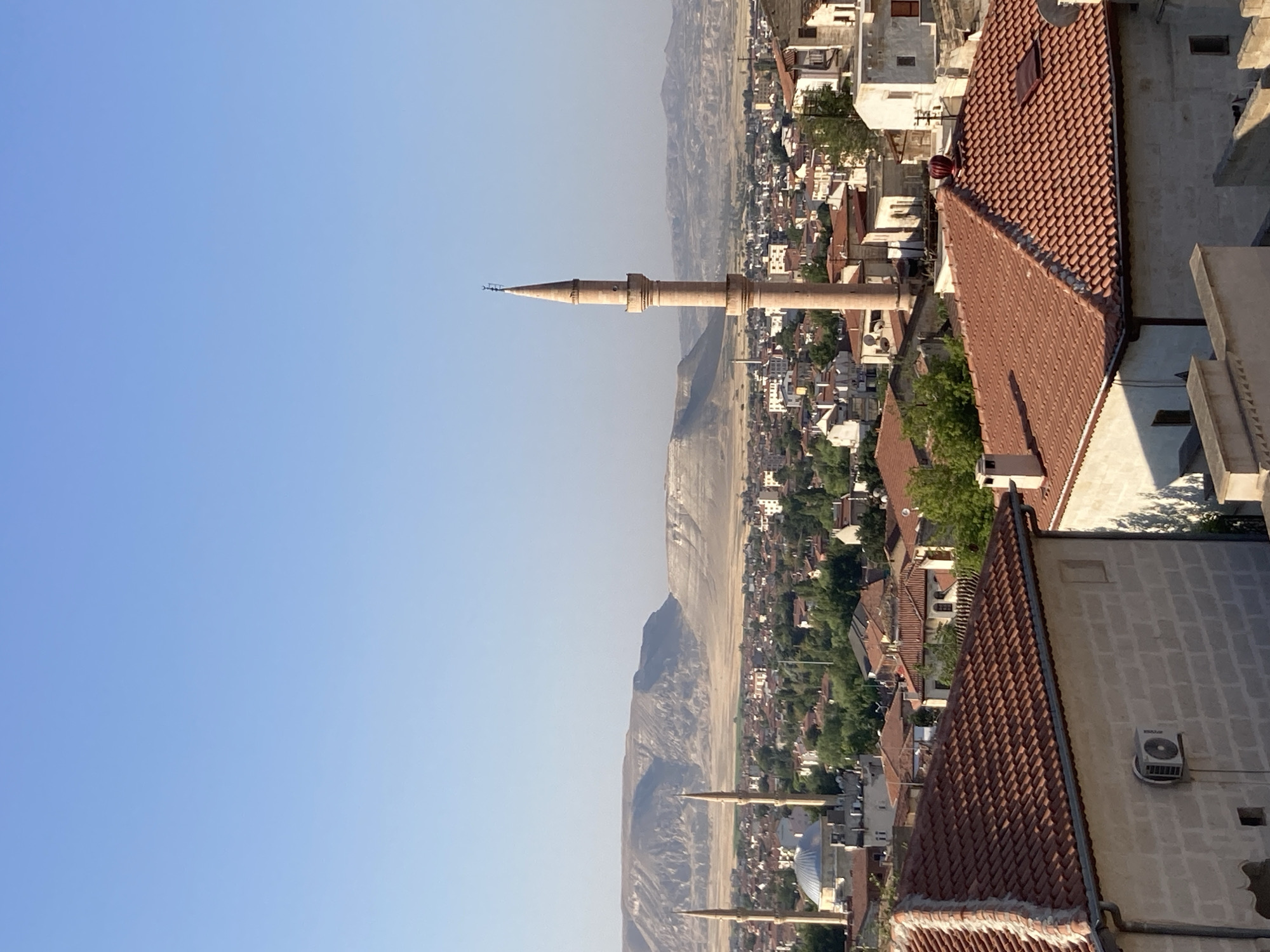 A panoramic view of Avanos İlçe Merkezi, Turkey, under a clear blue sky. The foreground features rooftops of traditional Turkish houses, characterized by their red-tiled surfaces and light-colored stone or brick walls. An air conditioning unit is visible on one of the walls. In the middle ground, the city unfolds with a dense cluster of buildings, interspersed with greenery. Two prominent minarets rise above the cityscape, flanking a mosque with a domed roof. Further in the distance, a long, arid mountain range stretches across the horizon, its layered rock formations creating a stark and rugged landscape. The overall scene suggests a calm, sunny day in a historic town nestled amidst a natural, mountainous environment.
