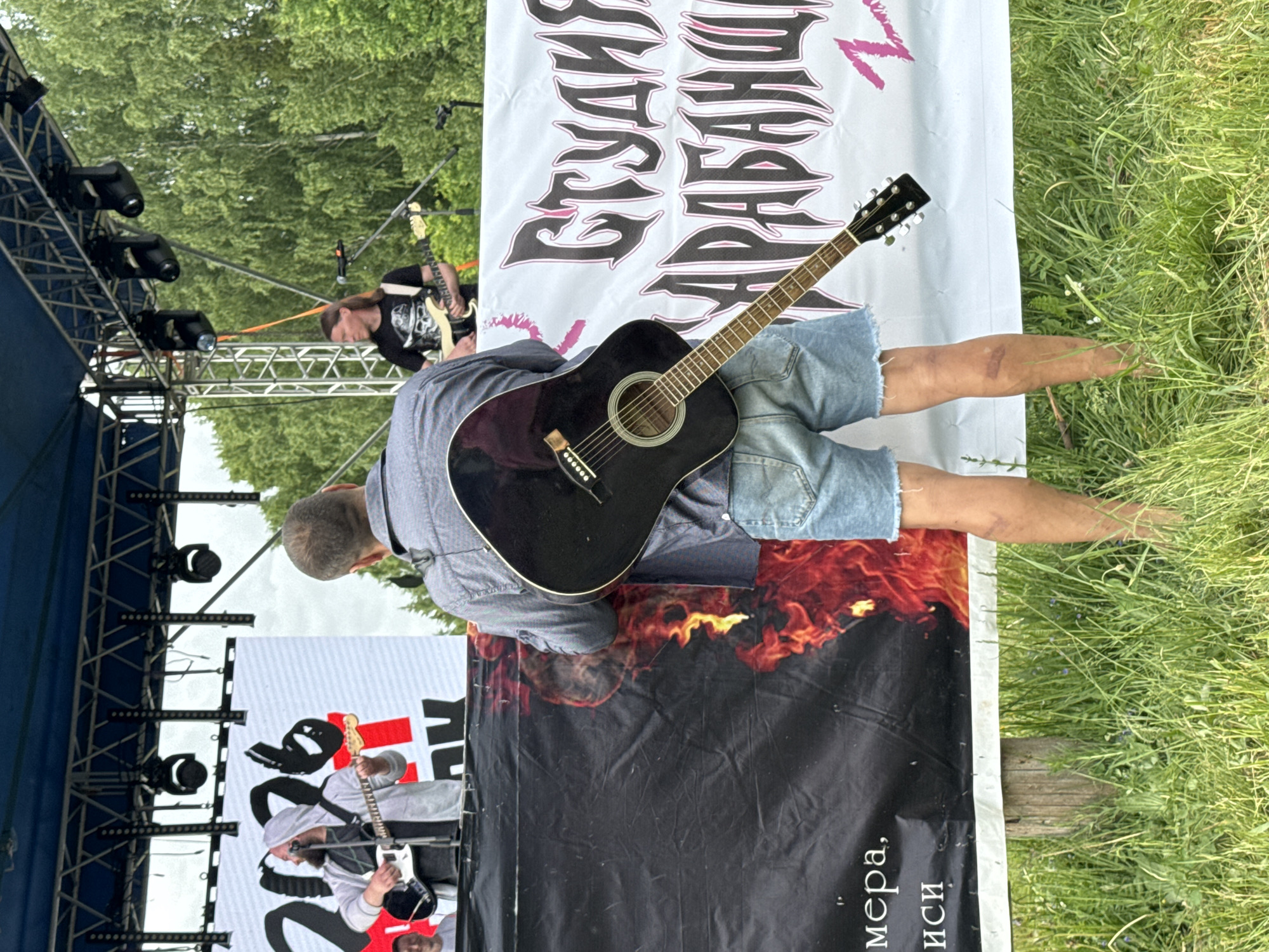 This image captures an outdoor music event, likely a concert or festival, during daylight hours. In the foreground, a person is seen from behind, lying or sitting on a large banner on the ground. They are wearing a blue or grey shirt and denim shorts, with a black acoustic guitar resting across their body.
On a stage positioned to the left (when viewing the image upright), two male musicians are actively performing. The musician closer to the camera wears a light-colored hooded top, glasses, and a beard, playing a light-colored electric guitar. Further back on the stage, another musician with long dark hair is playing an electric guitar, dressed in a dark shirt with a skull graphic. The stage is constructed with metal trussing and equipped with multiple black spotlights.
Several large banners are visible. The person in the foreground is resting on a black banner featuring a prominent red and orange flame graphic at the bottom. Partial white Cyrillic text can be seen on this banner, including words like "мера" and "иси." Behind this person, a white banner displays large black and pink stylized Cyrillic text that clearly reads "СТУДИЯ БАРАБАНОВ" (Studio of Drums). Another white banner with black and red graphics is partially visible to the left of the stage structure.
The setting is an open, grassy area surrounded by green trees, characteristic of an outdoor festival or park venue. The weather appears fair. The overall scene conveys the atmosphere of a live music performance in a relaxed outdoor environment.
