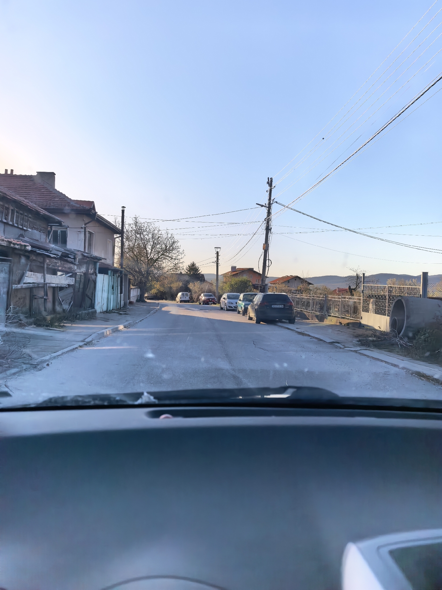 The image shows a view from inside a car, looking out onto a residential street in Unknown, Unknown. Several parked cars are visible along the right side of the street. Houses and other buildings line the street, with utility poles and power lines overhead. In the distance, the landscape appears hilly. The road surface is light-colored, possibly concrete or asphalt, and the sky is pale blue. The car's dashboard is visible in the foreground.
