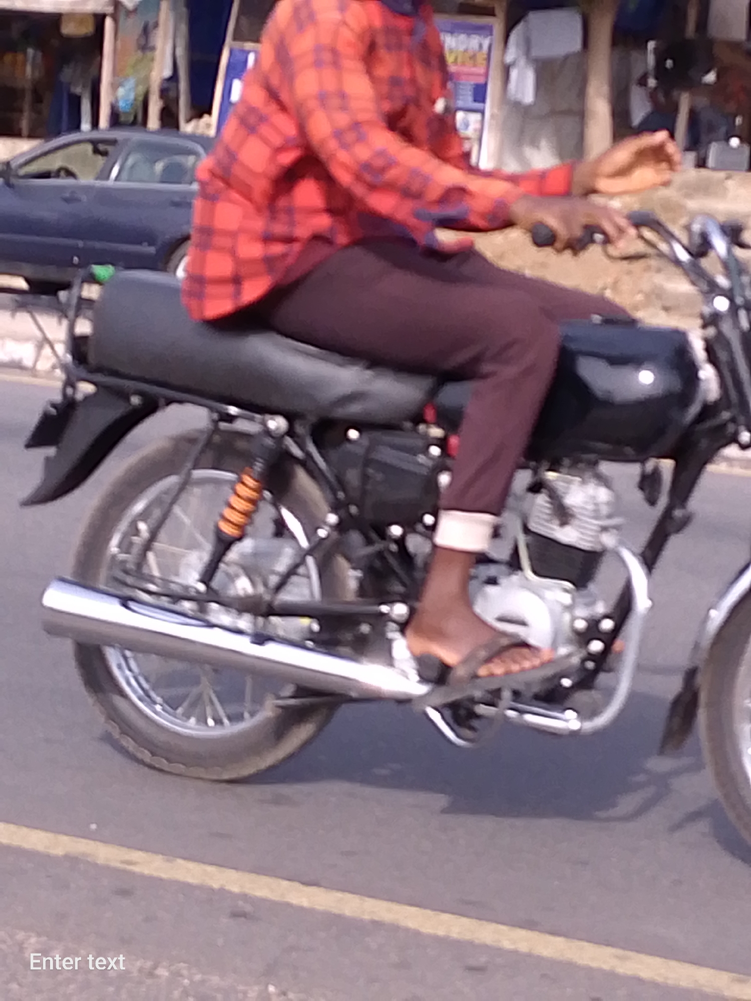 A person wearing a red checkered shirt and maroon pants is riding a motorcycle on a paved road. The motorcycle has a black gas tank and seat, with chrome exhaust and fenders. The rider is wearing sandals. A dark blue car is parked on the side of the road. The background includes buildings and other vehicles, suggesting an urban environment, specifically in Adankolo, Nigeria.