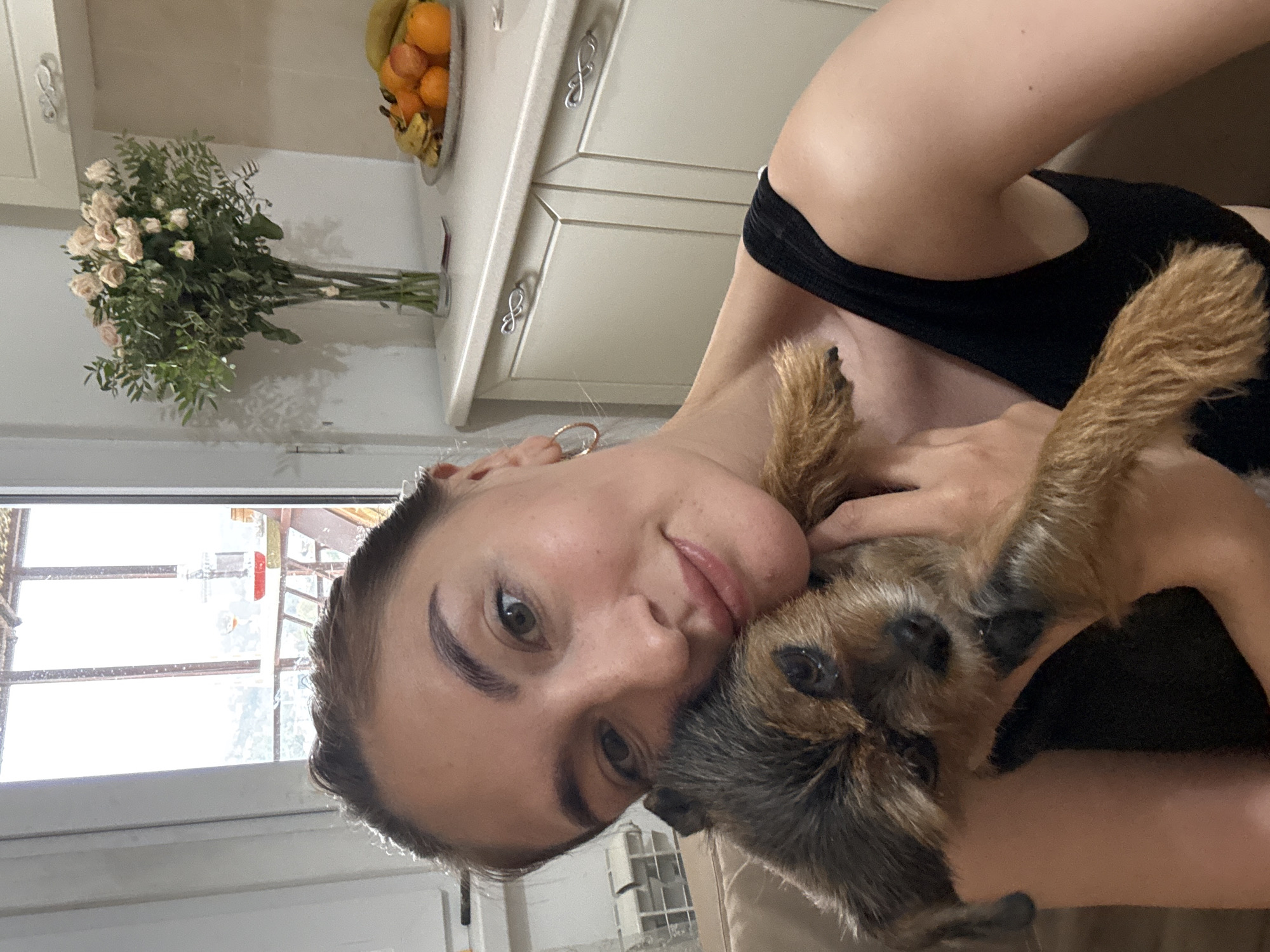 The image, oriented horizontally, presents a warm and intimate close-up of a young woman embracing a small dog in an indoor setting, likely a kitchen or adjacent living space in Sochi, Russia.
The central subjects are a young woman and a small, scruffy-haired dog. The woman has fair skin, dark, well-defined eyebrows, and light-colored eyes. Her dark hair is pulled back, and she wears a black top. She looks directly at the camera with a gentle, soft smile, her face tilted towards the dog.
The dog, appearing to be a terrier breed with brown and dark fur, is nestled affectionately against the woman's cheek. It also gazes towards the camera, appearing calm and comfortable in her arms.
The background, though slightly out of focus, reveals elements of a domestic environment. Light-colored kitchen cabinets with decorative, infinity-loop-shaped handles are visible. On a countertop, a clear glass vase holds a bouquet of light-hued roses (possibly pale pink or white) and green foliage. Nearby, a bowl brimmed with an assortment of fruits, including oranges/tangerines and bananas, sits on the counter. A large window provides bright natural light, suggesting it is daytime. Through the window, some external structures, possibly part of a building or scaffolding, can be vaguely discerned.
The image captures a tender moment of affection and companionship between the woman and her pet. No visible text is present in the scene.