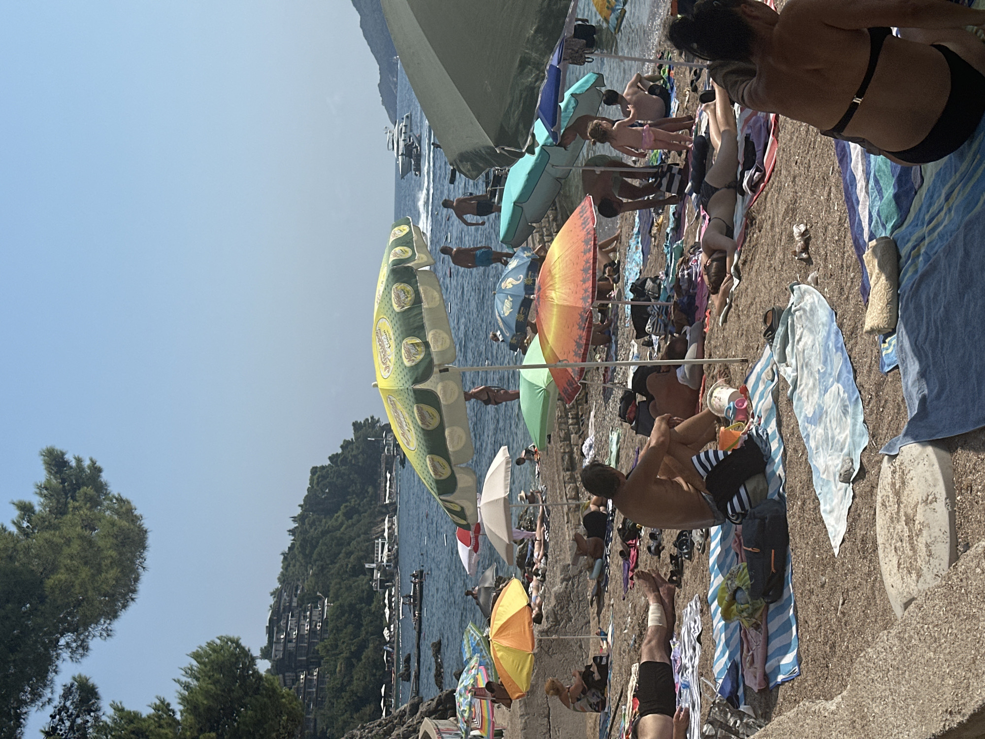 The image depicts a crowded beach scene in Budva, Montenegro, on a sunny day. A variety of beachgoers are scattered across the pebble beach, many relaxing under colorful umbrellas. Several individuals are sunbathing on towels, while others are standing or walking. In the background, the blue Adriatic Sea is visible, with a few boats, including a white yacht, cruising on the water. The coastline is lined with lush green hills, and buildings are visible on the slopes, suggesting the presence of a town or resort area. Umbrellas of various colors and sizes, towels, bags, and beach toys are strewn across the sand, indicating a typical beach day with leisure activities. The overall atmosphere appears to be one of a busy tourist destination.