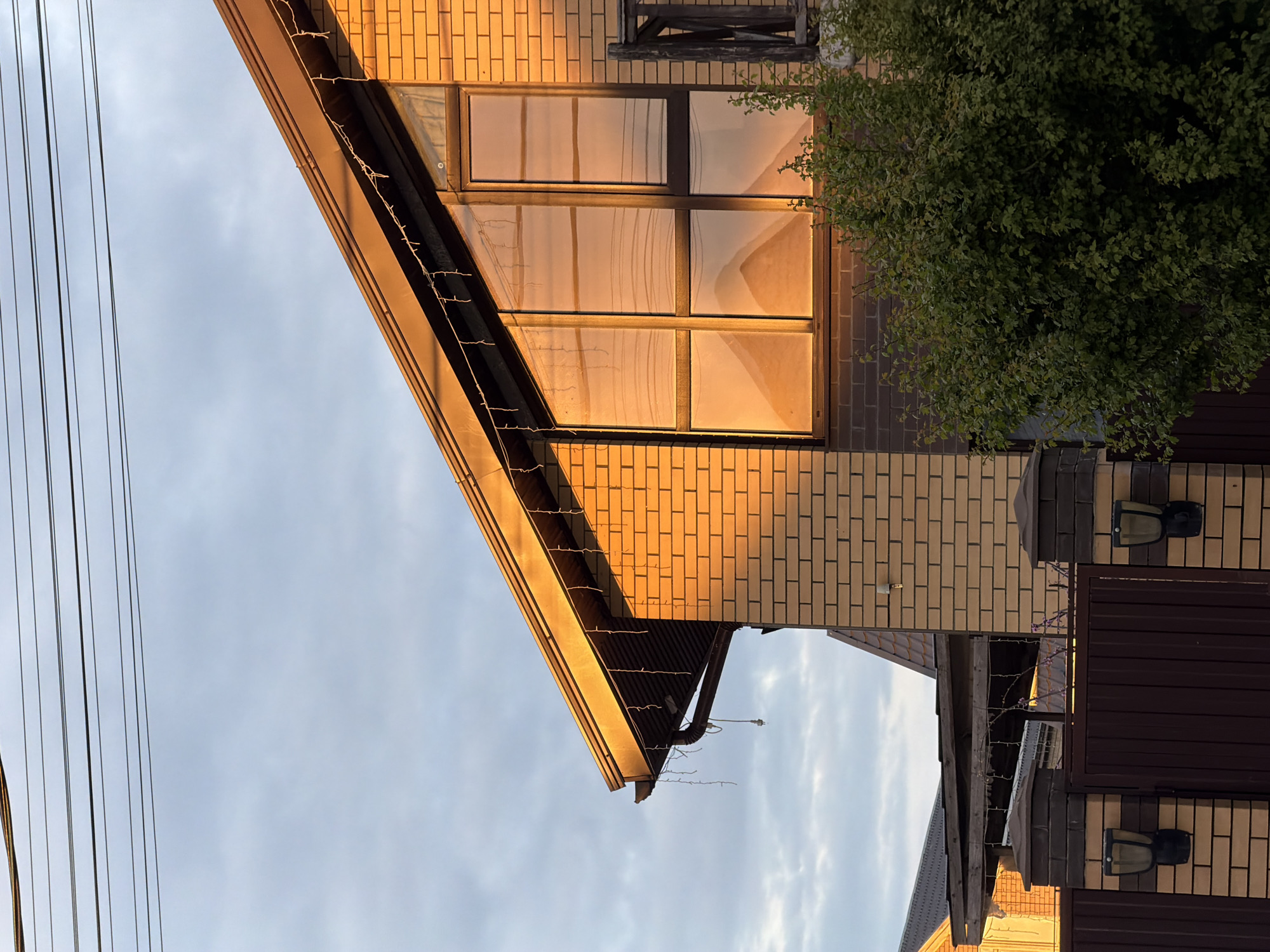 The image depicts a view of a building facade, likely a house, with brick walls and a gable roof. The windows are reflecting a warm, golden light, suggesting either sunrise or sunset. Some wires run diagonally across the sky in the upper left corner, possibly electrical or telephone lines. To the right of the building, a small section of a fence and a tree are visible. The scene appears to be located in a residential area. There are no visible people, specific actions, or interactions in the frame. The location is Rossiyskiy, Russia.