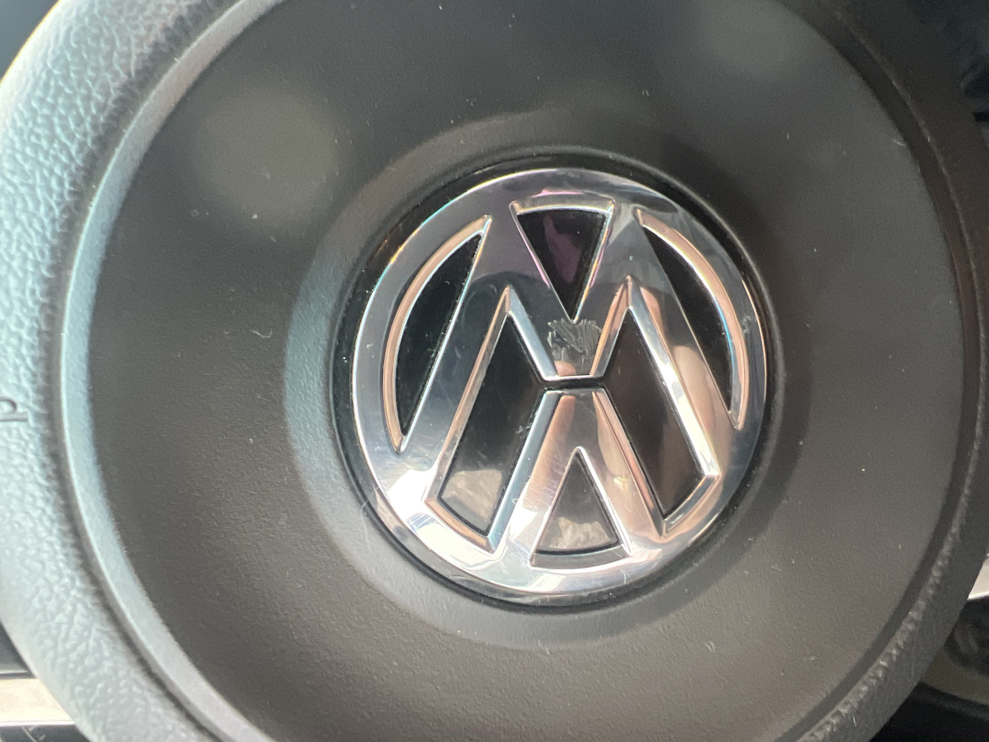 A close-up, overhead shot captures the Volkswagen logo centered on a black steering wheel. The logo, a silver metallic emblem, features the letters "V" and "W" superimposed within a circle. The steering wheel's textured surface and a subtle sheen suggest it is made of plastic and is in a vehicle. The lighting indicates an indoor setting, likely within a car, with reflections visible on the logo. The background is a dark, textured material consistent with a car's interior. The image does not provide any information or context related to Yanino-1, Russia, or any people.
