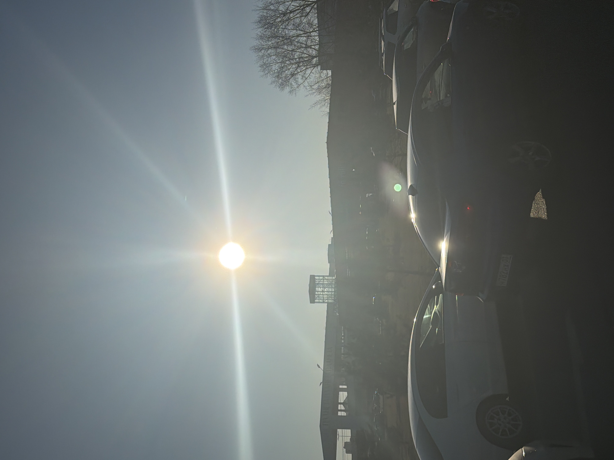 The image is a heavily backlit, outdoor scene in what appears to be an urban area. The sun is a prominent feature, positioned relatively high in the sky, creating significant lens flare and making details difficult to discern. A row of parked cars is visible to the right, heavily obscured by the sun's glare. A structure resembling a building or industrial facility can be seen in the background. The low contrast and bright light make specific details like street signs or the presence of people unclear. A leafless tree is silhouetted against the sky. Given the license plate on the car, this may be located in Krasnoyarsk, Russia.