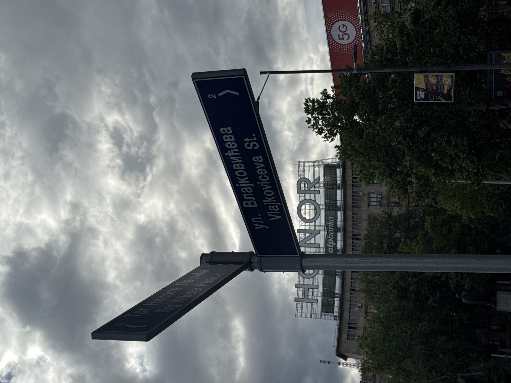 The image shows a street sign in Belgrade, Serbia, indicating the intersection of Trg Nikole Pasica and Vlajkoviceva Street. The sign for Vlajkoviceva Street also displays the number "2" and an arrow pointing in that direction. In the background, there's a building with a large "HINOR" sign and the logo for "otpbanka" displayed on it. There are also trees and a 5G advertisement in the background. The sky is overcast with gray clouds.
