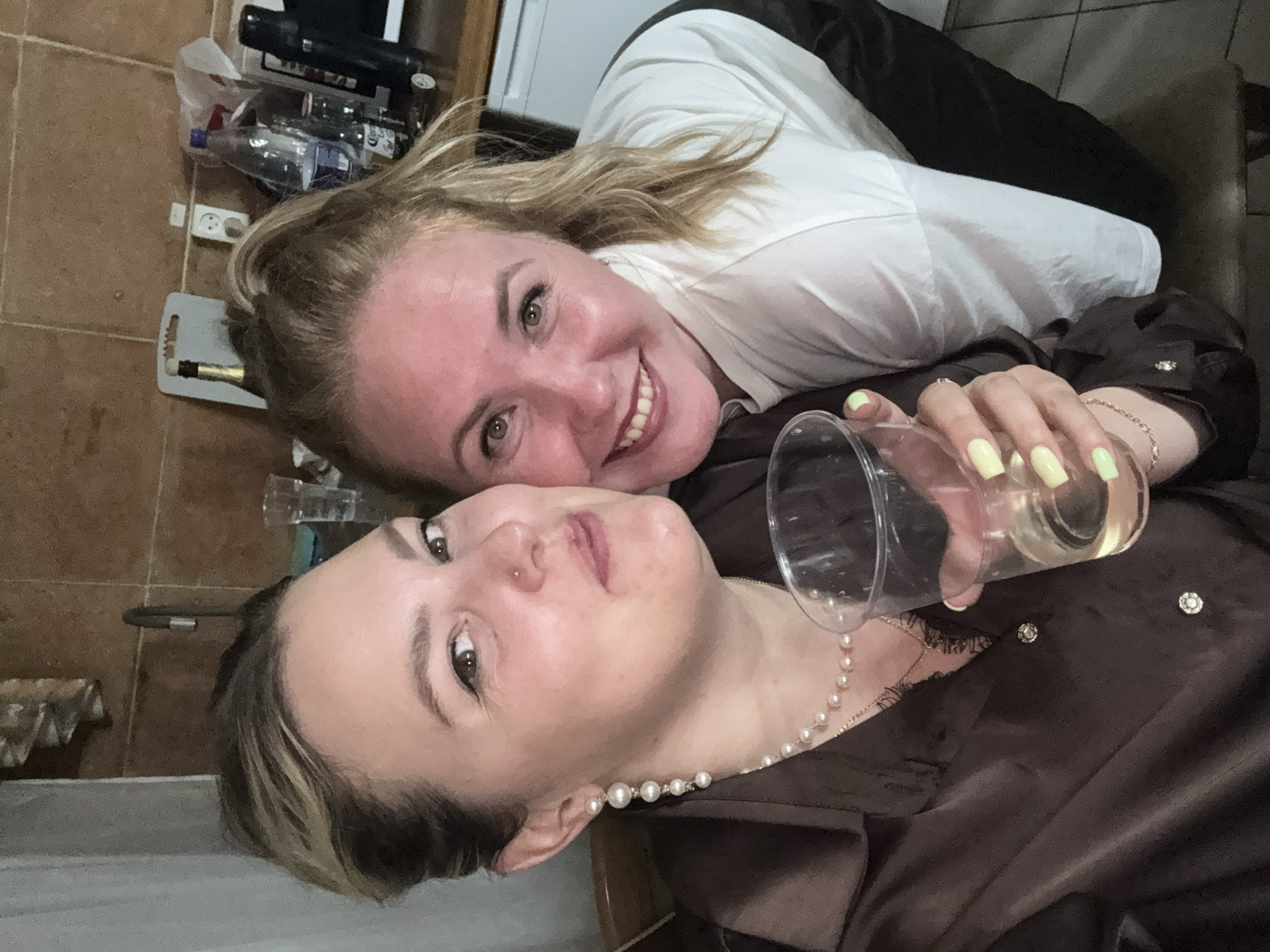 Two women are taking a selfie in what appears to be a kitchen area. The woman on the left is wearing a brown satin shirt with pearl earrings and a pearl necklace. She is holding a clear plastic cup with a light-colored liquid. The woman on the right is wearing a white t-shirt and has her blonde hair pulled into a ponytail. Both women are smiling and looking at the camera. In the background, there are kitchen cabinets, a sink, a bottle of Corona beer, and other miscellaneous items on a countertop. The lighting suggests it is indoors, possibly in the evening. The scene is consistent with a casual gathering or party. There is no information in the image to suggest the location is Zavod Mikhailovskii, Russia.
