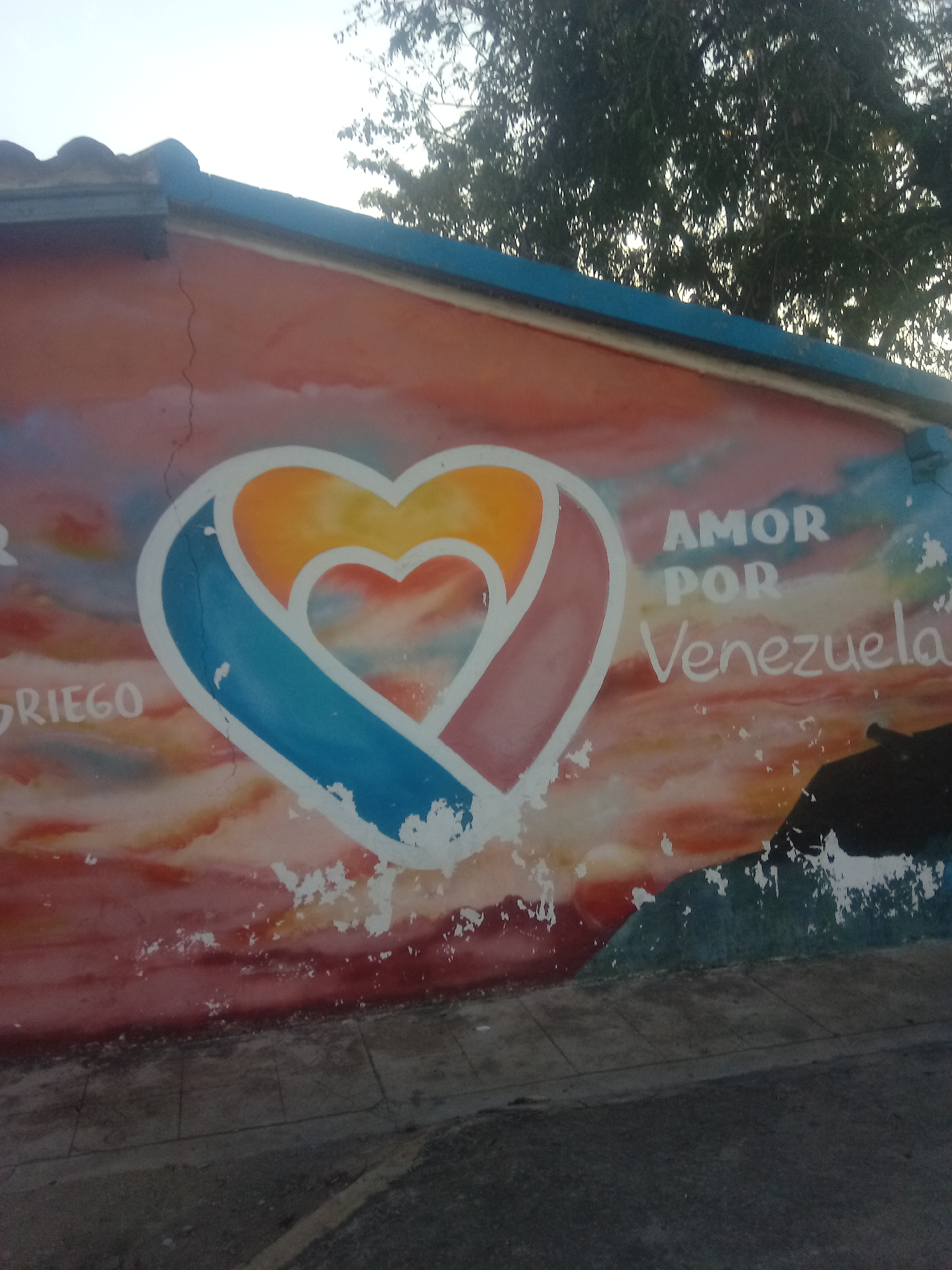 The image shows a mural on a building in Juan Griego, Venezuela. The mural depicts three concentric hearts, with the inner heart in red, the middle heart in orange, and the outer heart in blue and pink. Next to the hearts are the words "Amor Por Venezuela" in white letters. The mural is painted on a weathered wall with a blue trim. The background of the mural is a blend of colors resembling a sunset. Part of a tree is visible in the top portion of the image. The bottom shows the edge of a paved area.