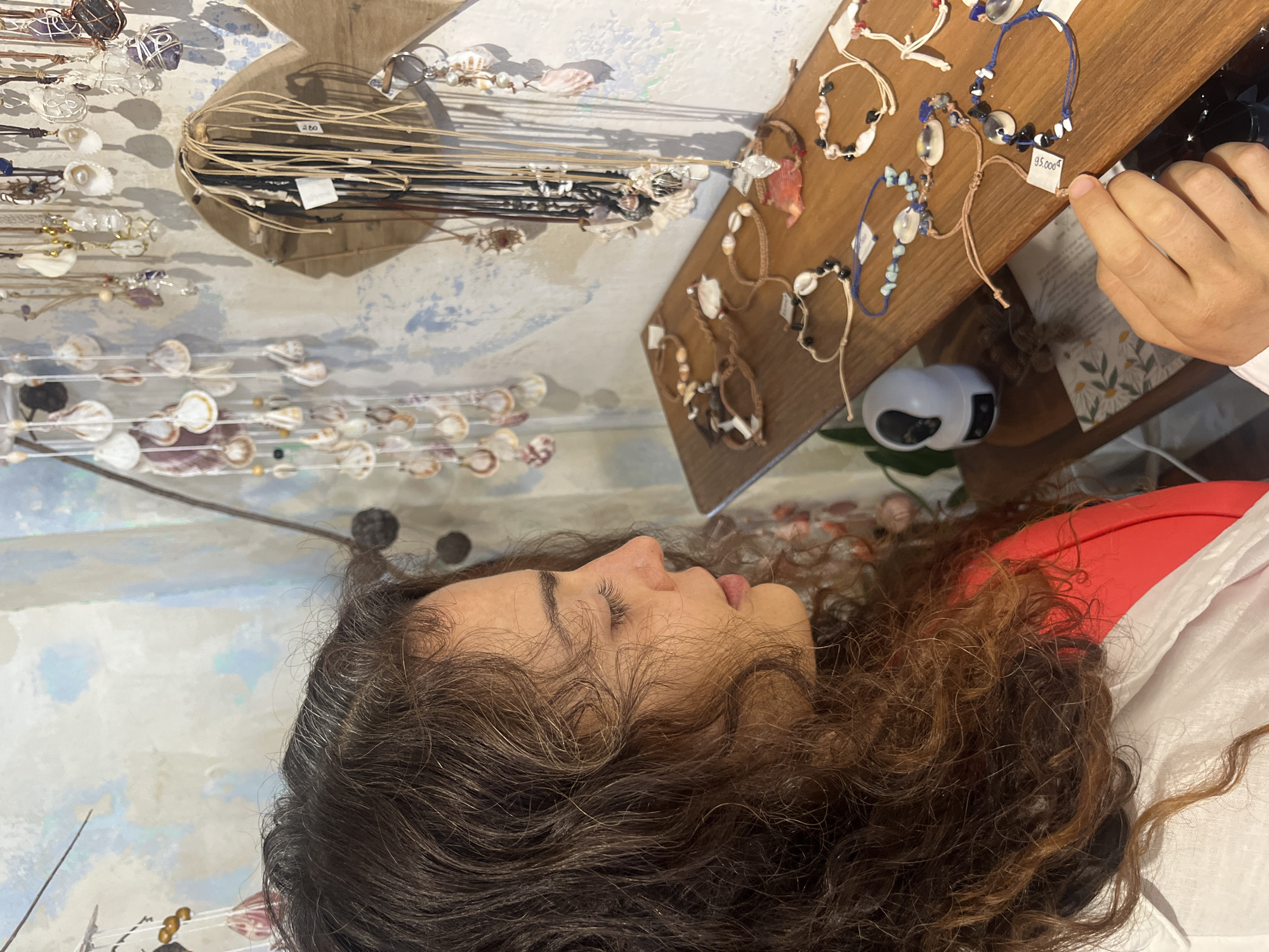 A young woman with dark, curly hair is shown in a side profile, looking at a display of handmade jewelry. She is wearing a white shirt over a red garment. The jewelry, primarily necklaces and bracelets, features shells, stones, and beads, and is displayed on a wooden shelf and hanging on a wall adorned with more shell decorations. A small security camera is visible on the shelf. The background suggests an indoor environment, possibly a shop, with a textured, painted wall. The overall scene appears to be in a market or boutique setting in Phu Thuy Ward, Vietnam, indicated by the presence of handcrafted items and the implied location.
