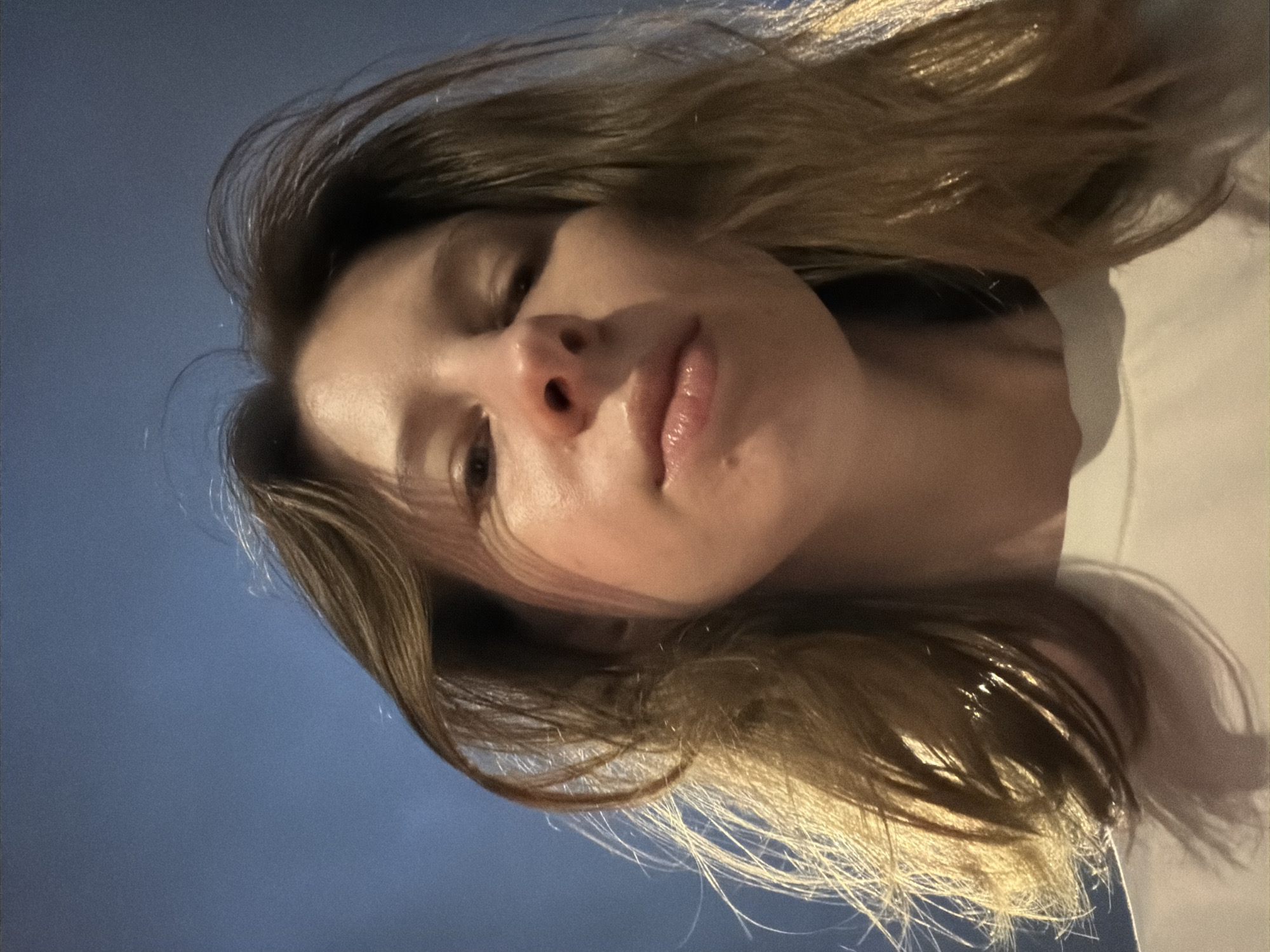 A low-angle, close-up shot shows the face of a young woman against a dark blue sky, suggesting it was taken outdoors at night. The woman has light brown hair that is windswept around her face, and her skin is illuminated by a warm light source, casting shadows on her features. She is wearing a white t-shirt. The camera is positioned directly in front of her, looking slightly upwards. The image does not contain any discernible landmarks or features that would indicate a specific location within Dongducheon-si, South Korea.