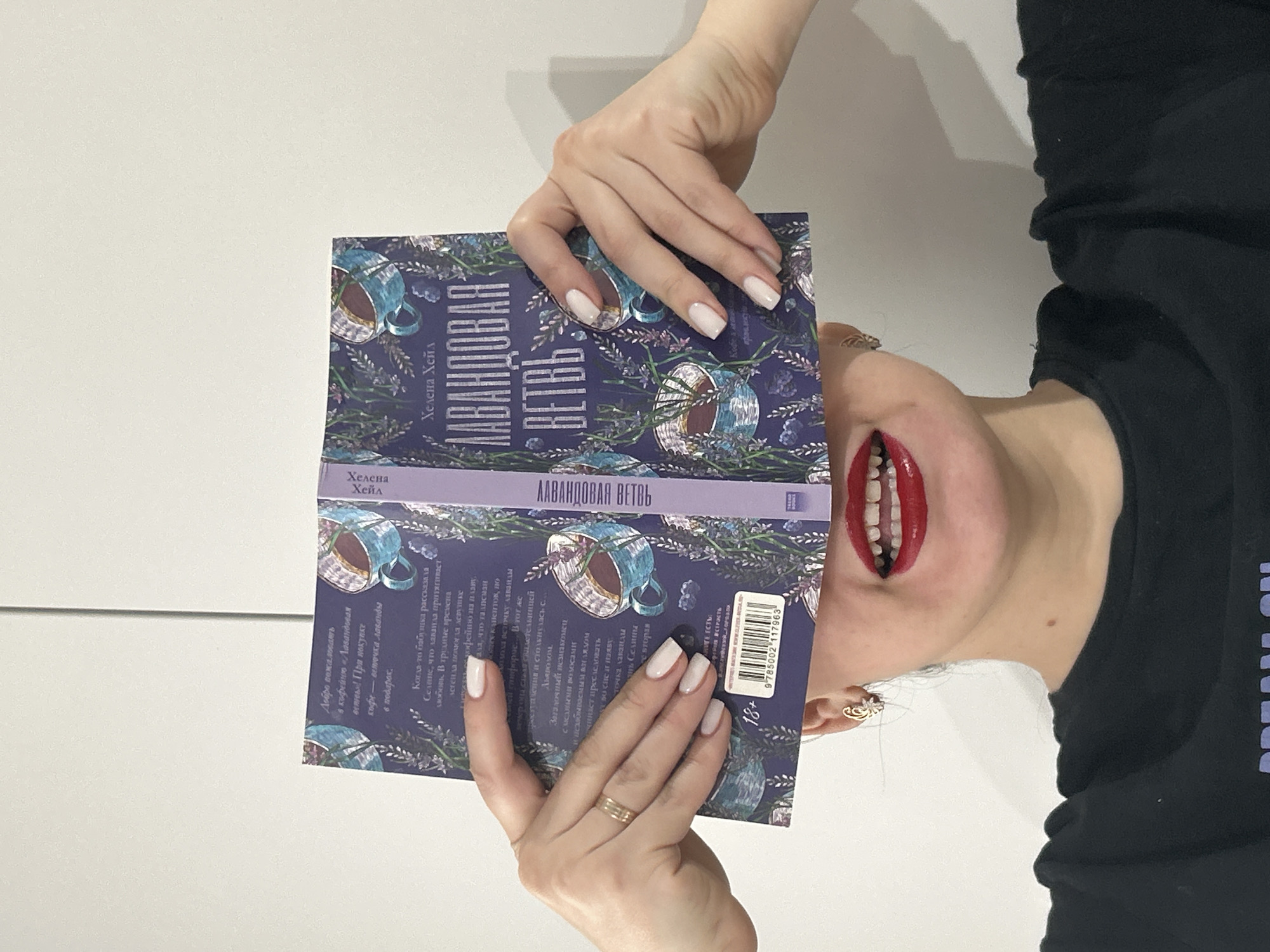 The image, taken in Chaikovsky, Russia, is rotated 90 degrees counter-clockwise and depicts a person, likely a woman, holding a book. The background is a plain, light-colored wall or surface.
The person's hands, adorned with light-colored manicured nails, hold the book open. A gold-toned ring is visible on the left ring finger, and a decorative earring is seen on the right earlobe. The person's face is partially visible, showing a chin and mouth with bright red lipstick. The mouth is open, revealing teeth, suggesting a stylized or playful expression. The person is wearing a black top, with the words "DREAM ON" partially visible on the right side of the frame.
The book's title, clearly visible on the front and spine, is "ЛАВАНДОВАЯ ВЕТВЬ" (Lavender Branch/Twig), authored by "Хелена Хейл" (Helena Hale). The cover features illustrations of lavender sprigs and teacups. The back cover provides more details, mentioning a coffee shop called "Лавандовая ветвь" that offers a lavender twig as a gift with coffee. It also references a story about lavender attracting love, an "18+" age rating, and a barcode with ISBN "9785002117963". The scene is set indoors, with no specific time of day or weather conditions evident.