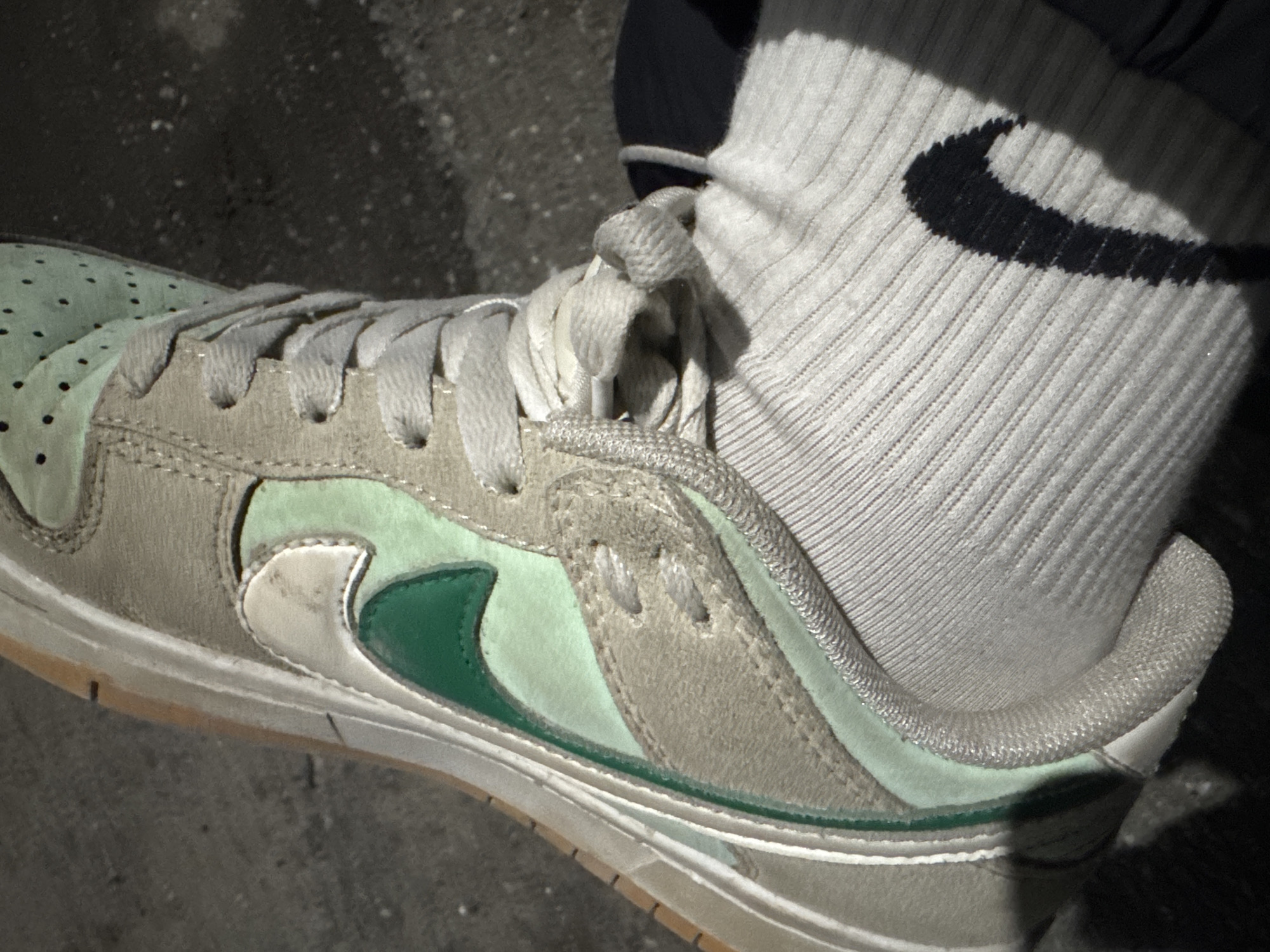 A close-up shot of a sneaker on a foot wearing a white sock. The sneaker is primarily gray suede with light green panels and a dark green Nike swoosh. The sock is a ribbed crew sock with a black Nike swoosh visible. The location is undiscernible from the image and the presence of the city Yerevan, Armenia cannot be confirmed.