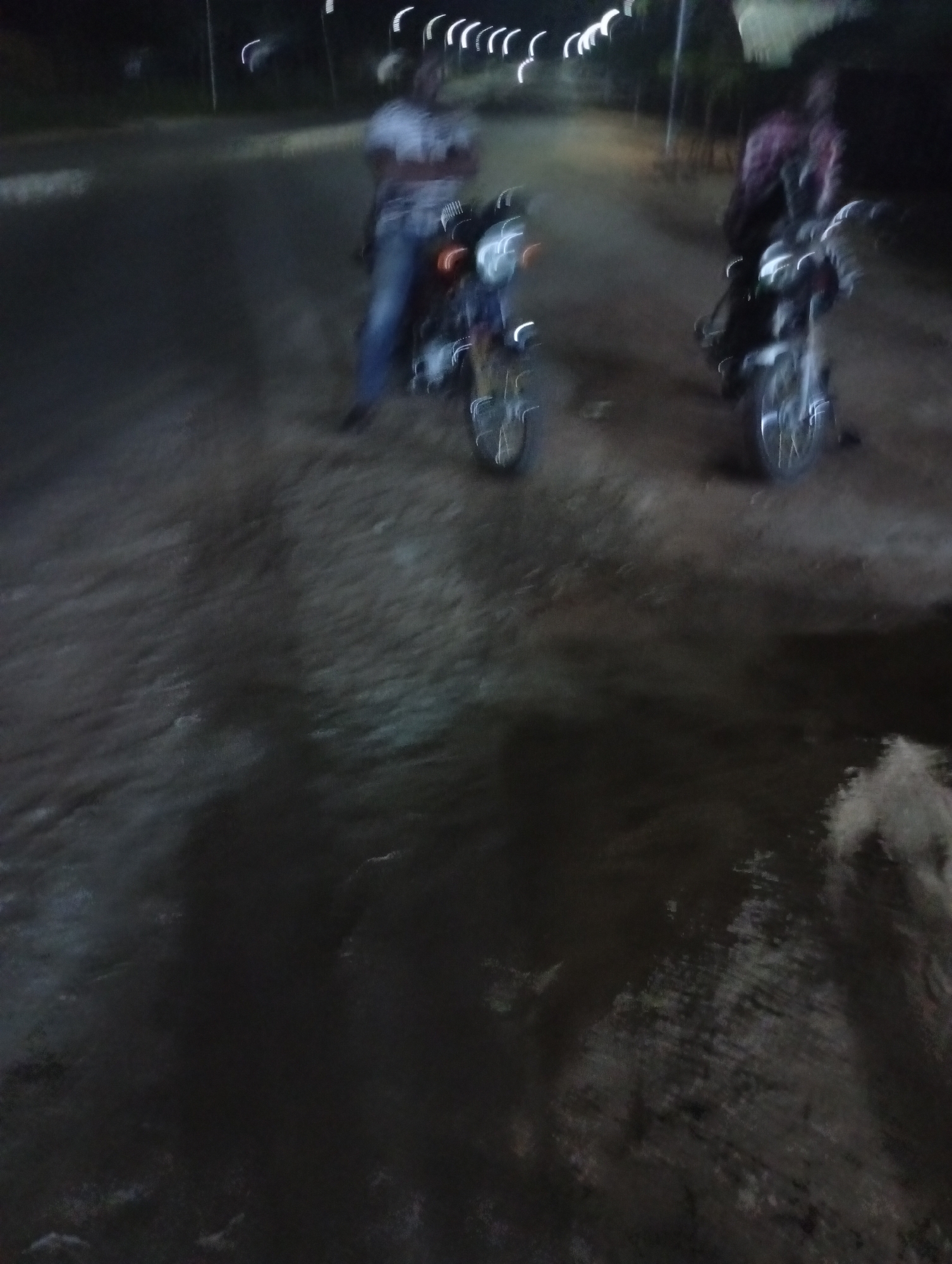 The image depicts a dimly lit street in Adankolo, Nigeria, at night. Two motorcycles are parked, each with a person standing nearby. The person on the left wears light-colored clothing and blue jeans, while the person on the right wears a darker, patterned top. The road surface appears uneven and possibly unpaved. Overhead streetlights create blurry streaks in the sky.