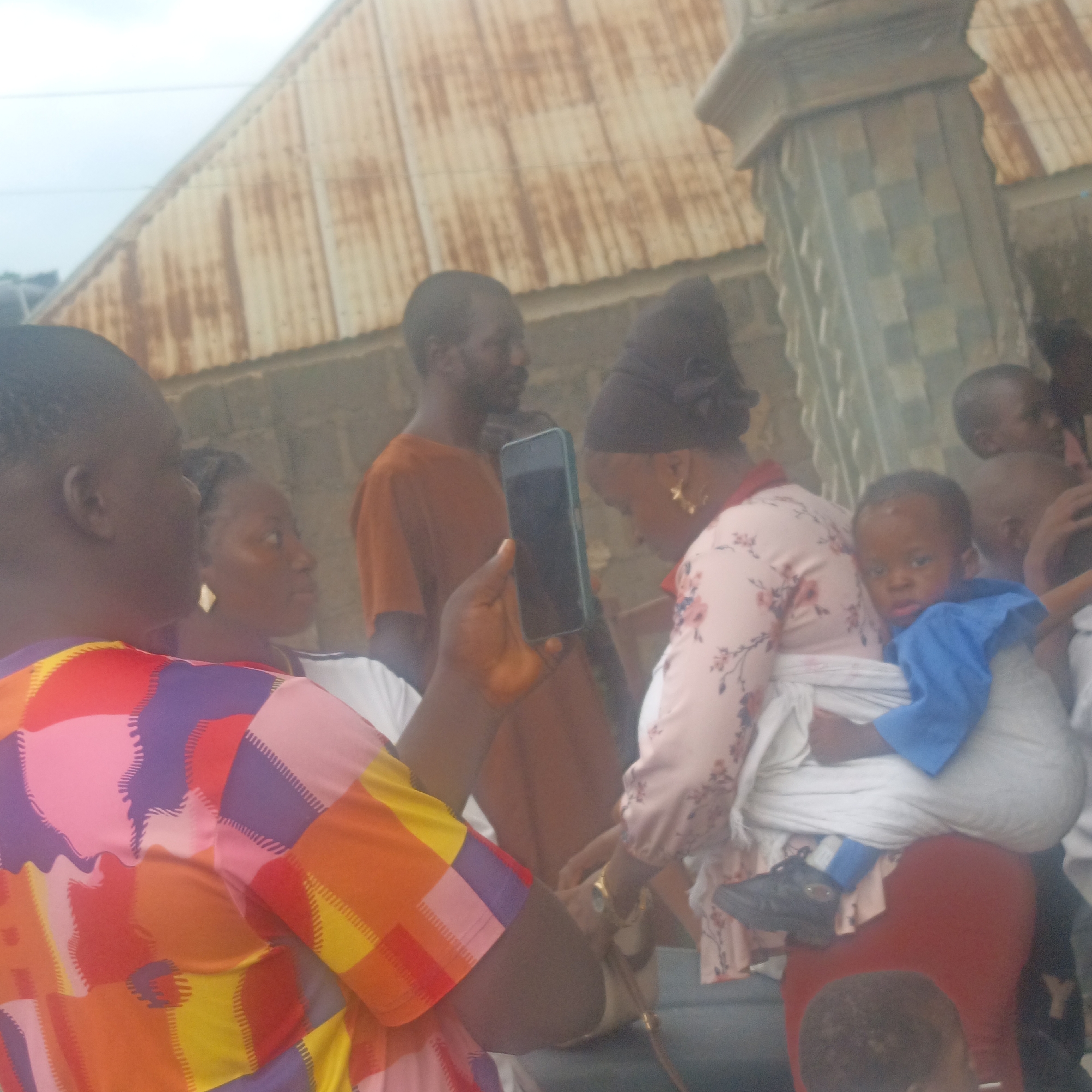This is a medium-shot, eye-level, outdoor photograph taken during the day in Akwanga, Nigeria. The image captures a group of people, with a primary focus on several individuals in the foreground and mid-ground.
In the immediate foreground, a person wearing a brightly colored, multi-patterned shirt (pink, purple, red, orange, yellow) is seen from the back and side. Their left arm is raised, holding a dark-colored smartphone in a green case.
To the right of this individual, a woman is visible from the side, wearing a pink floral-patterned top and a headscarf. She is holding a baby wrapped in a white cloth and a blue garment. The baby, with dark skin and hair, is facing towards the camera and appears to be around one to two years old. The woman's face is partially obscured by the baby and her headscarf. She is wearing large, gold hoop earrings.
Behind the woman and the baby, a man in an orange-brown shirt is standing and looking to the left. To the far left of the frame, another person's dark head is visible in profile.
In the background, a corrugated metal roof with visible rust stains is the dominant element, suggesting a simple, possibly semi-rural or informal urban setting. A weathered, textured wall runs horizontally behind the people. To the far right, a section of a decorative concrete pillar or column is visible. The sky above is bright and slightly hazy. The overall impression is one of a community gathering or an everyday street scene.