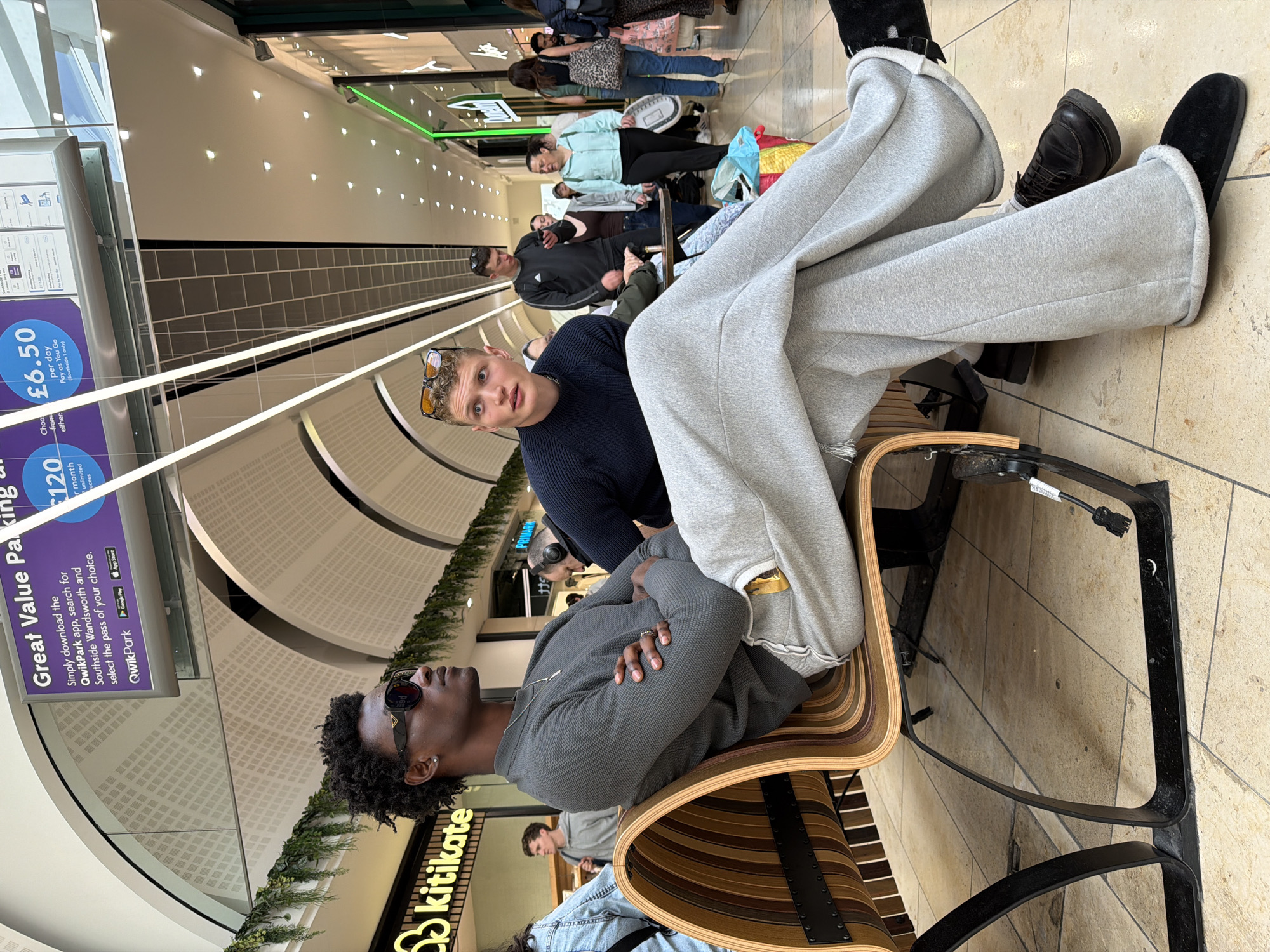 The image shows an indoor scene, likely a shopping mall in Southside Wandsworth, London, United Kingdom. Two people are sitting on chairs, one is wearing sunglasses and a grey sweater, with his arms crossed. Another person is wearing a blue sweater and sunglasses on his forehead, looking at the camera. There are other people walking in the background. The mall appears to have a curved ceiling with decorative greenery. Signs for parking services, including "QwikPark" and prices, are visible. A "kitikate" store sign is also present.