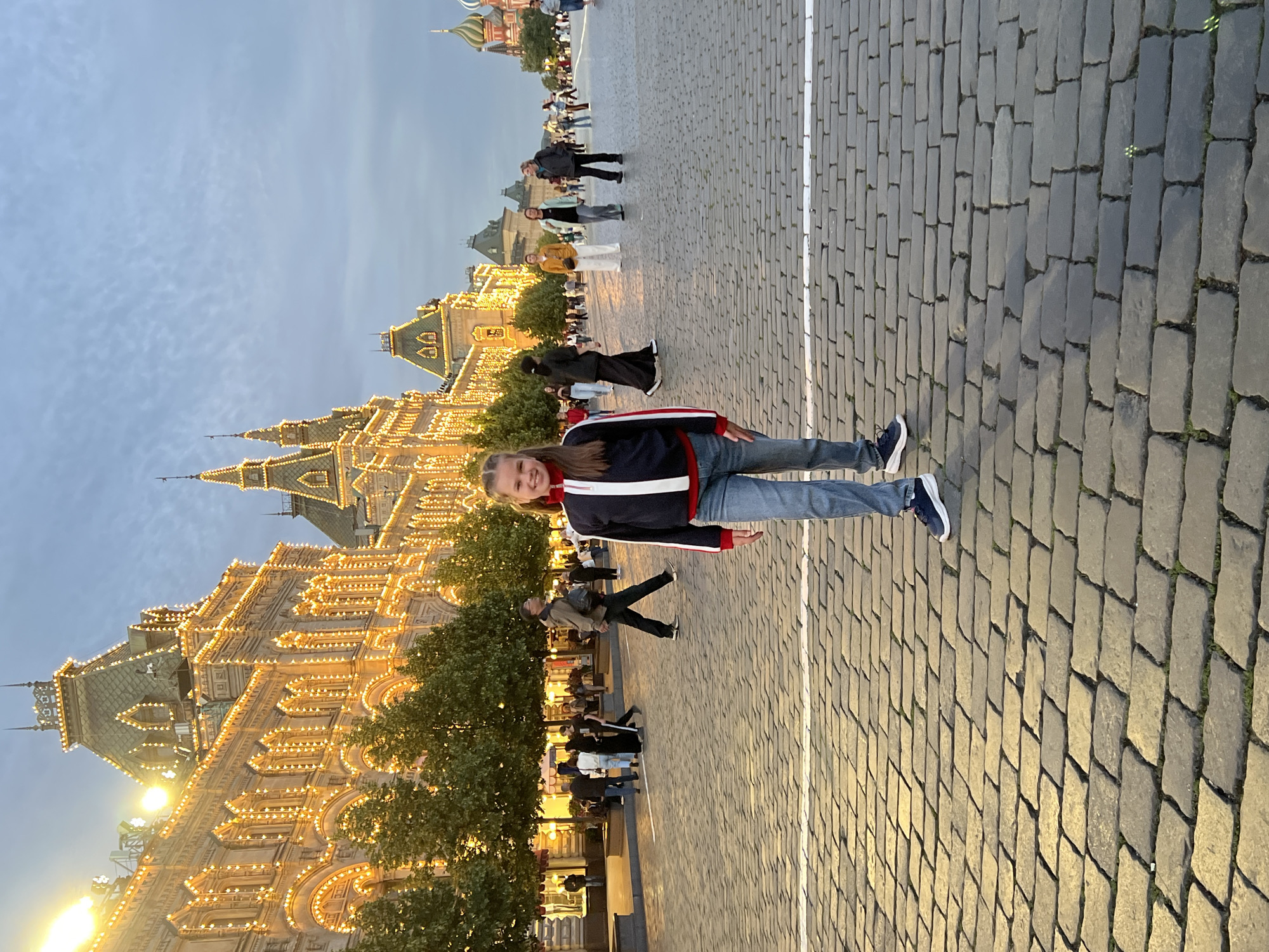 The image depicts a young girl standing on a cobblestone square in Moscow, Russia. The girl is positioned in the foreground, facing the camera with a smile. She is wearing a navy blue jacket with red and white stripes, light blue jeans, and dark sneakers. Behind her, several people are walking on the square, and in the background, a grand building with ornate architecture and numerous lights is visible. The sky above is a twilight blue, suggesting the time is late evening or early morning. The overall scene appears to be Red Square, a prominent historical and cultural site in Moscow, known for its iconic architecture and vast open space.