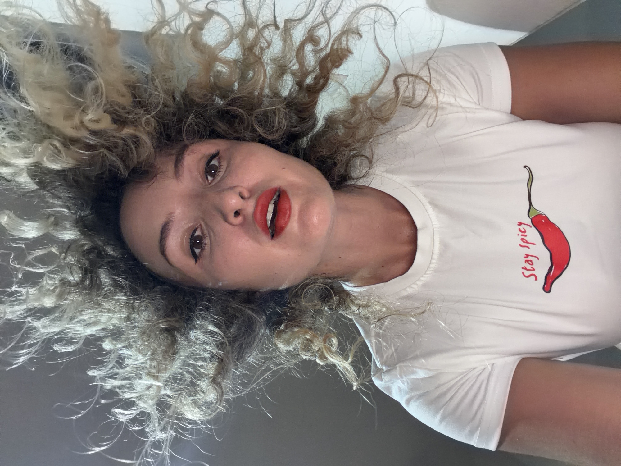 The media file is a close-up, low-angle portrait of a young woman lying down, with her head tilted back. She has curly, light brown and blonde hair that fans out around her head. Her face is illuminated, and she is wearing bright red lipstick and eyeliner. Her eyes are open and looking towards the camera.
She is wearing a white t-shirt with a graphic of a red chili pepper and the text "Stay Spicy" written in red to the right of the pepper. The setting appears to be indoors, with a white background and a greyish surface below. The lighting suggests it might be daytime. The woman's expression is playful and engaging.
The orientation of the image is such that the woman's head is at the top and her body extends downwards. This perspective, combined with the direct gaze and bright makeup, creates an intimate and striking image. There are no obvious cues about the weather or time of day beyond the general indoor lighting. The location context provided is Baan Bang Rak, Thailand, though no visual elements within the image specifically indicate this location.