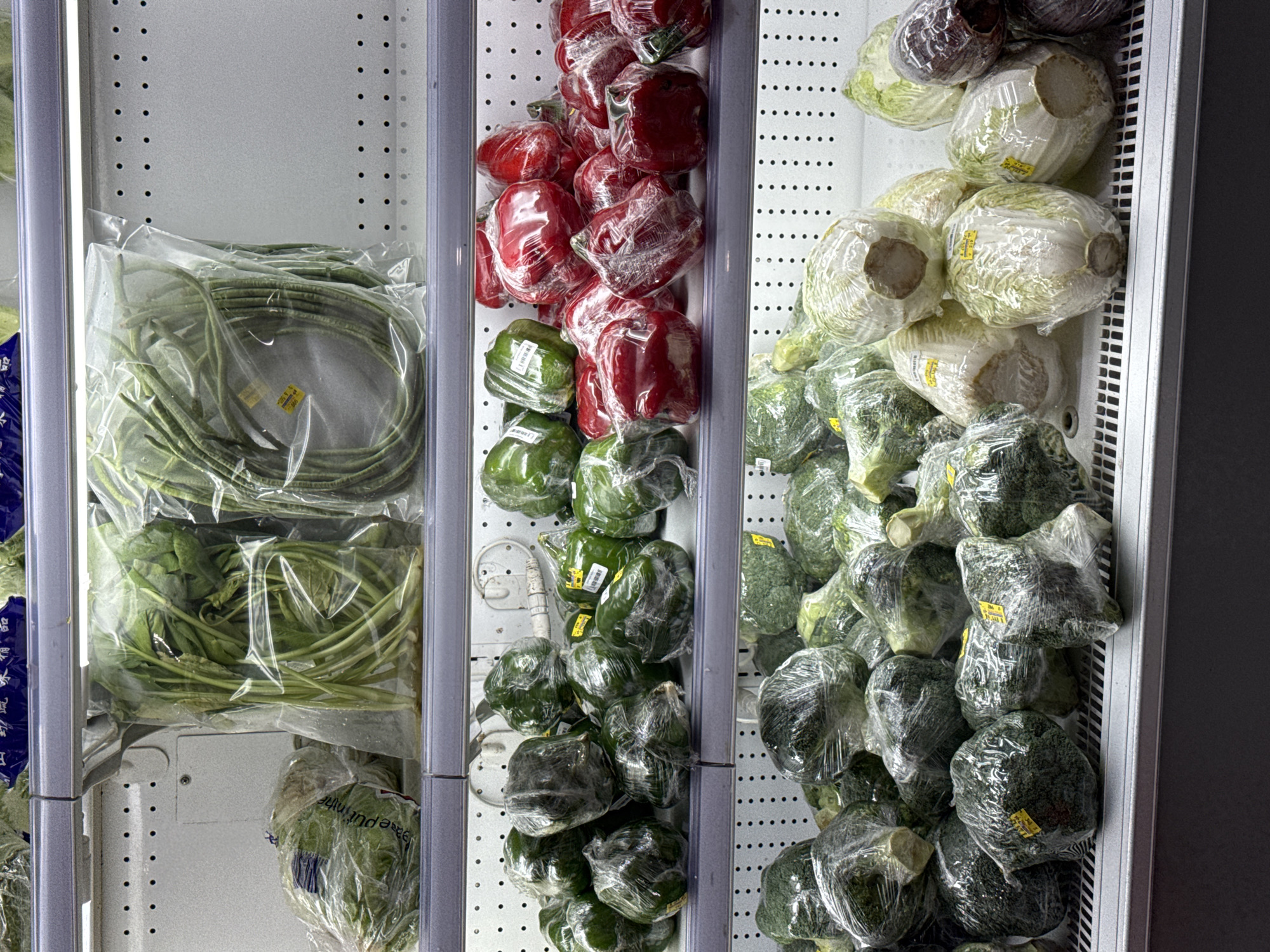 The image shows a refrigerated display case filled with various fresh vegetables in a market. On the top shelf, there are large bunches of leafy green vegetables wrapped in plastic, alongside long green beans also packaged in plastic. The middle shelf displays a variety of bell peppers, with green and red peppers individually wrapped in plastic. The bottom shelf is filled with broccoli florets and heads of napa cabbage, all similarly packaged. The display case appears to be in a well-lit environment, likely a grocery store or market. Based on the provided information, this scene is located in the city of Tanjung Uban, Indonesia.
