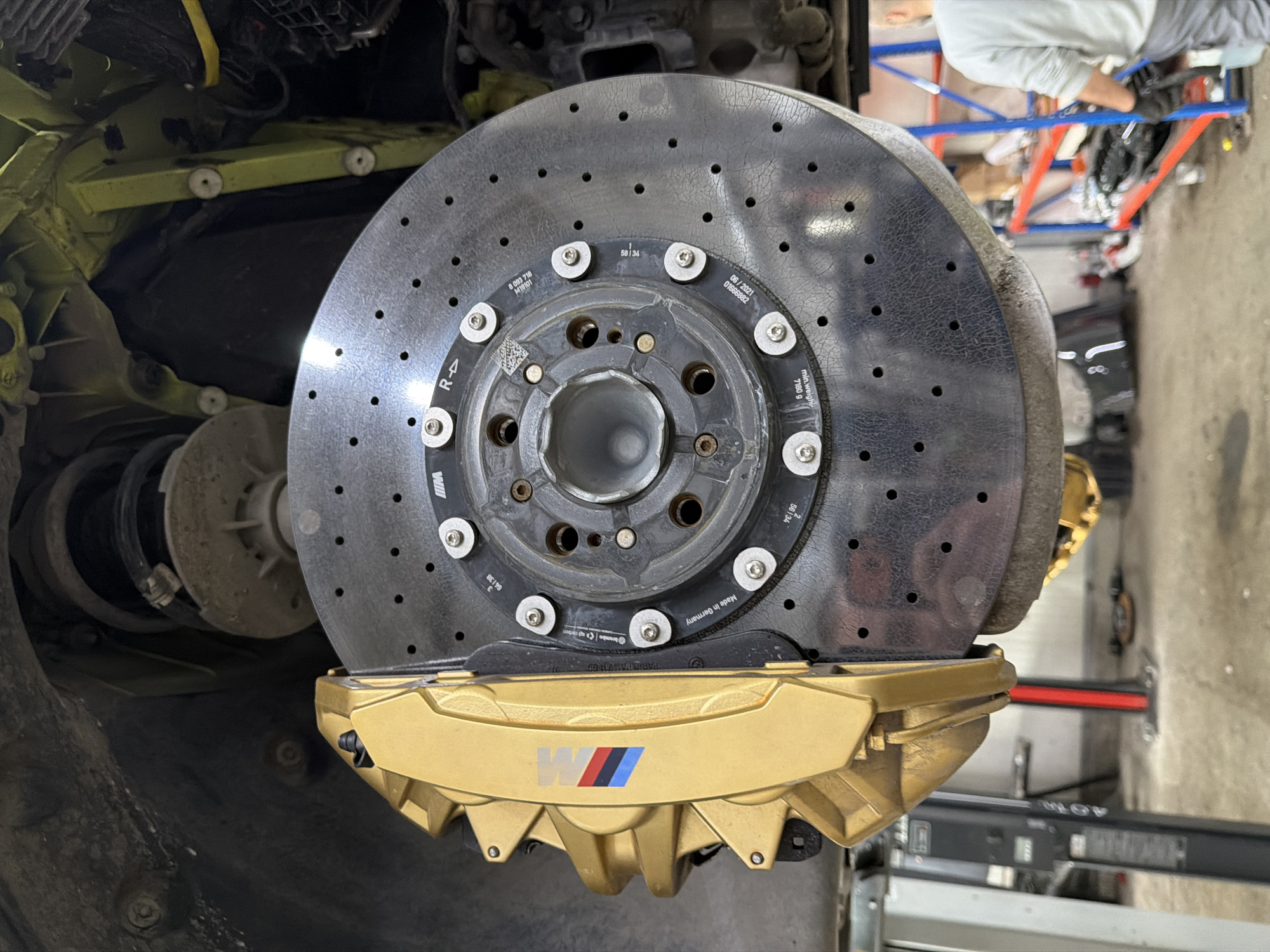 A close-up, rotated image shows a high-performance vehicle's braking system, prominently featuring a large, dark gray perforated carbon-ceramic brake rotor and a gold-colored multi-piston caliper. The rotor displays signs of use with slight cracking patterns on its surface and is attached to a central hub with multiple bolts. The gold caliper, positioned beneath the rotor, has the distinct red, dark blue, and light blue BMW M logo.
In the background, parts of the vehicle's suspension and chassis are visible, including a dirty, light green/yellow painted structural component and a coil spring assembly. The scene appears to be an indoor workshop or garage setting, indicated by glimpses of a concrete floor, red and blue shelving, and a person's lower body (wearing light clothing) in the upper right background, suggesting maintenance or repair work is underway. The image is oriented vertically, with the brake assembly appearing on its side.
Visible text on the brake rotor includes "R <-" indicating its orientation, a date "06/2021" likely for manufacturing, "min.weight 7180 g", "Made in Germany", and the brand "Brembo carbon". Other alphanumeric codes such as "0 093 718", "M19101", "58134", "01666882", "64138", and "56134" are also present.