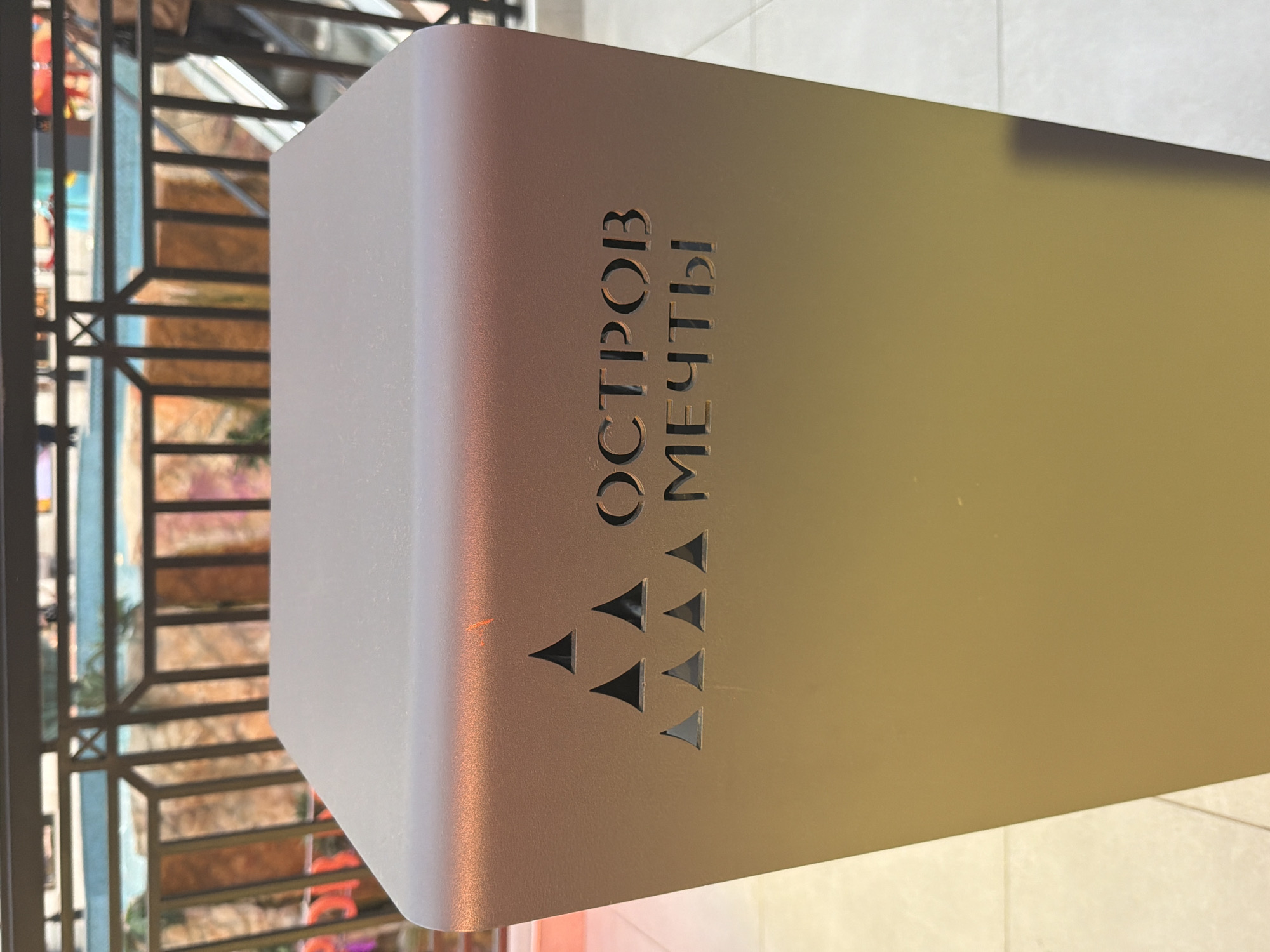 The image shows a close-up of a metallic pillar with text and geometric shapes carved into it. The pillar has a smooth, reflective surface with a gradient of colors, shifting from a muted pinkish-brown on the left to a yellowish-gold on the right, suggesting artificial lighting.
The text, written in Cyrillic letters, is oriented vertically and appears to be the name of a place or attraction. The first line reads "ОСТРОВ", and the second line reads "МЕЧТЫ". Below the text, there is a pattern of several small, triangular cutouts arranged in a staggered, upward-pointing formation. Some of these triangles are filled with a darker material, creating a contrast with the pillar's surface.
In the blurred background, a metal railing and a colorful, possibly indoor, environment with geometric patterns can be seen. The overall impression is that of a modern, possibly retail or entertainment, setting. There are no people visible in the image. The lighting suggests it might be daytime, but the indoor setting makes it difficult to determine. The mood is neutral, focusing on the design elements of the pillar.