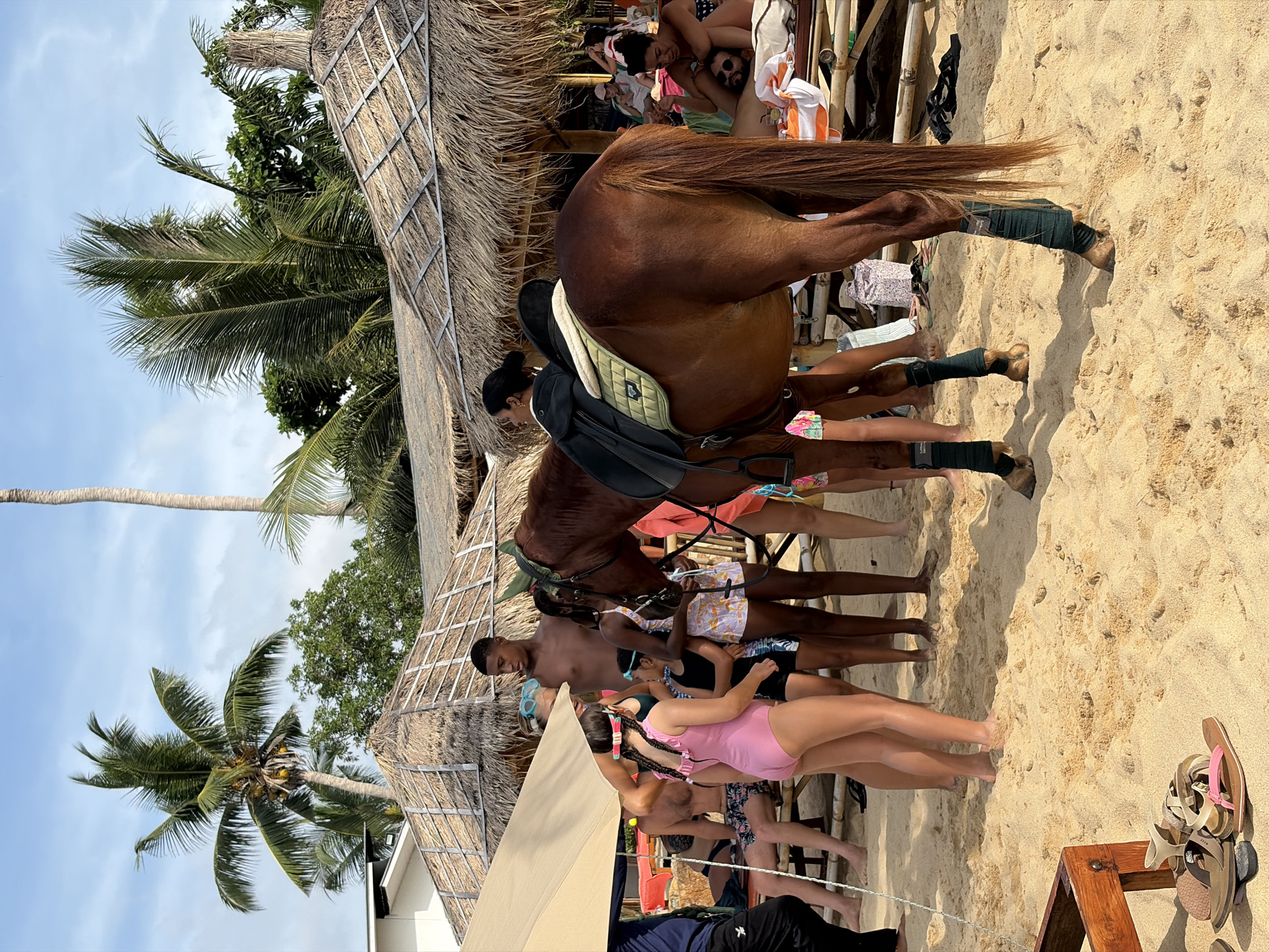 A brown horse is standing on a sandy beach in Ko Samui District, Thailand, facing right. The horse is equipped with a black saddle and a light green saddle pad. Dark green wraps are visible on its lower legs. Several people are gathered around the horse, with some children in the foreground. A young girl in a pink swimsuit is to the left of the horse, and another girl in a patterned dress is to the right. A tall young man is standing behind the horse, looking forward. In the background, there are thatched-roof structures, palm trees, and beach chairs, suggesting a resort or tourist area. Some individuals are visible relaxing on lounge chairs in the distance. The sky is partly cloudy. The sand is light brown and appears dry.