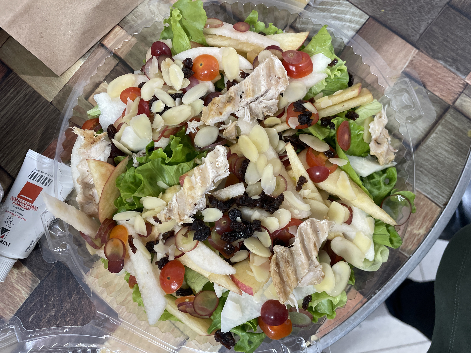The image displays a close-up, overhead view of a salad in a clear plastic container, positioned on a patterned surface resembling wood planks. The salad contains various ingredients, including leafy greens, sliced apples, cherry tomatoes, red grapes, sliced almonds, dried fruit (likely raisins), and pieces of grilled chicken. The container is transparent, allowing the contents to be clearly visible. In the upper left corner of the image, a white tube with red and black lettering is partially visible, suggesting it might be a medical or cosmetic product. The background shows a textured surface, with hints of a brown paper bag on the right side. The lighting suggests an indoor setting, possibly a restaurant or a food establishment. The image does not contain any discernible landmarks or contextual elements that would allow for pinpointing its exact location within Davao City, Philippines.