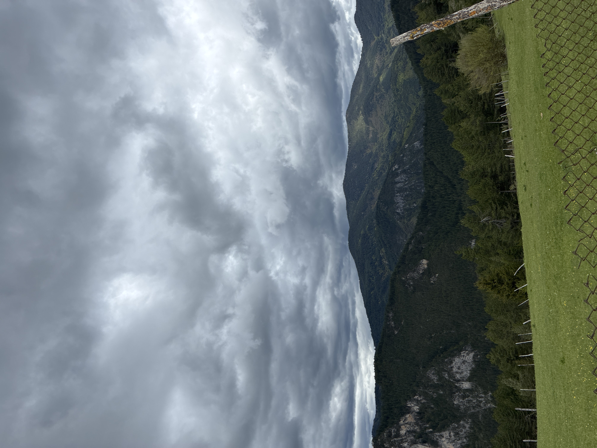The image shows a mountainous landscape under a cloudy sky. The photograph appears to be taken from a high vantage point, potentially near a meadow with a wire fence visible in the foreground. The mountains are densely forested. The sky is overcast with various cloud formations. The location is Giovren, Bulgaria.