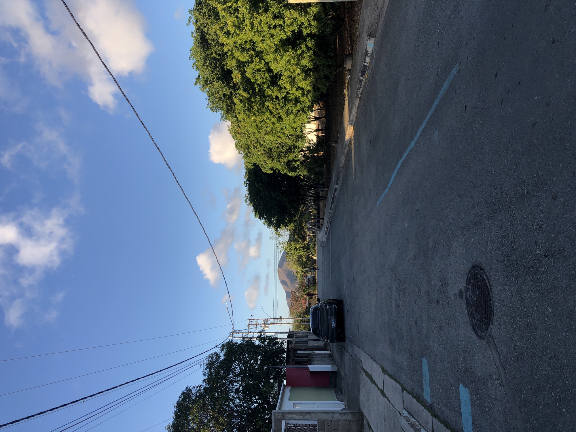The image shows a street scene in Altagracia, Venezuela. The road surface is asphalt with a blue painted line and a manhole cover on the right side. A dark-colored car is parked on the right side of the road, near buildings. Power lines stretch across the blue sky, which contains fluffy white clouds. Trees line the left side of the road. In the distance, a mountain is visible. There are people walking on the street in the distance. Some buildings can be observed on the left side of the road, some with a painted facade.