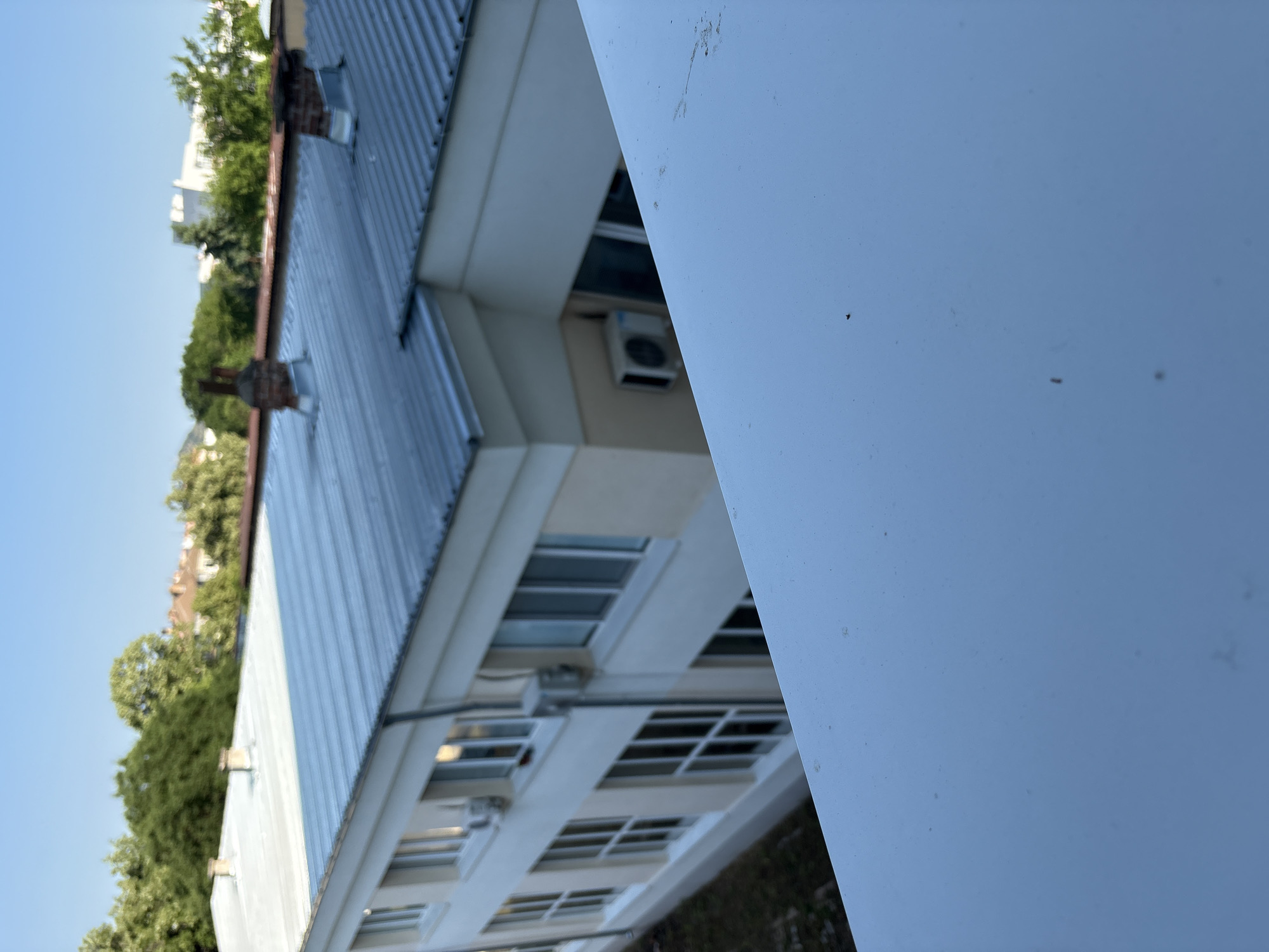 The image shows a view of a building with a metal roof, likely located in Plovdiv, Bulgaria. The building is light-colored, possibly cream or beige, with multiple windows. There are two chimneys on the roof, one made of brick. Trees and other buildings are visible in the background. The foreground is dominated by a white surface, possibly a railing or a balcony, which is out of focus. An air conditioning unit is mounted on the side of the building. The sky is clear and blue, indicating a sunny day.