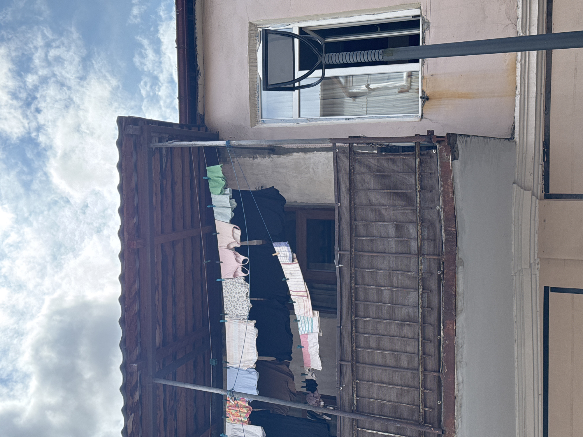 A low-angle, exterior shot captures a portion of a light pink building facade under a cloudy sky. A wooden-tiled balcony roof juts out from the building, supported by a metal pole. Clothes are hung on a line stretched across the balcony, secured by clothespins. The clothing includes various items such as shirts, underwear, and towels, in a range of colors and patterns. To the right of the balcony, a tall, dark grey street lamp stands in front of a window with white framing. The window is partially open, revealing a light-colored curtain inside. The building's facade exhibits some wear, with a rust-colored stain extending downwards from the window. The lower part of the image shows a concrete ledge and further architectural details of the building. The overall scene is situated in Batumi, Georgia.