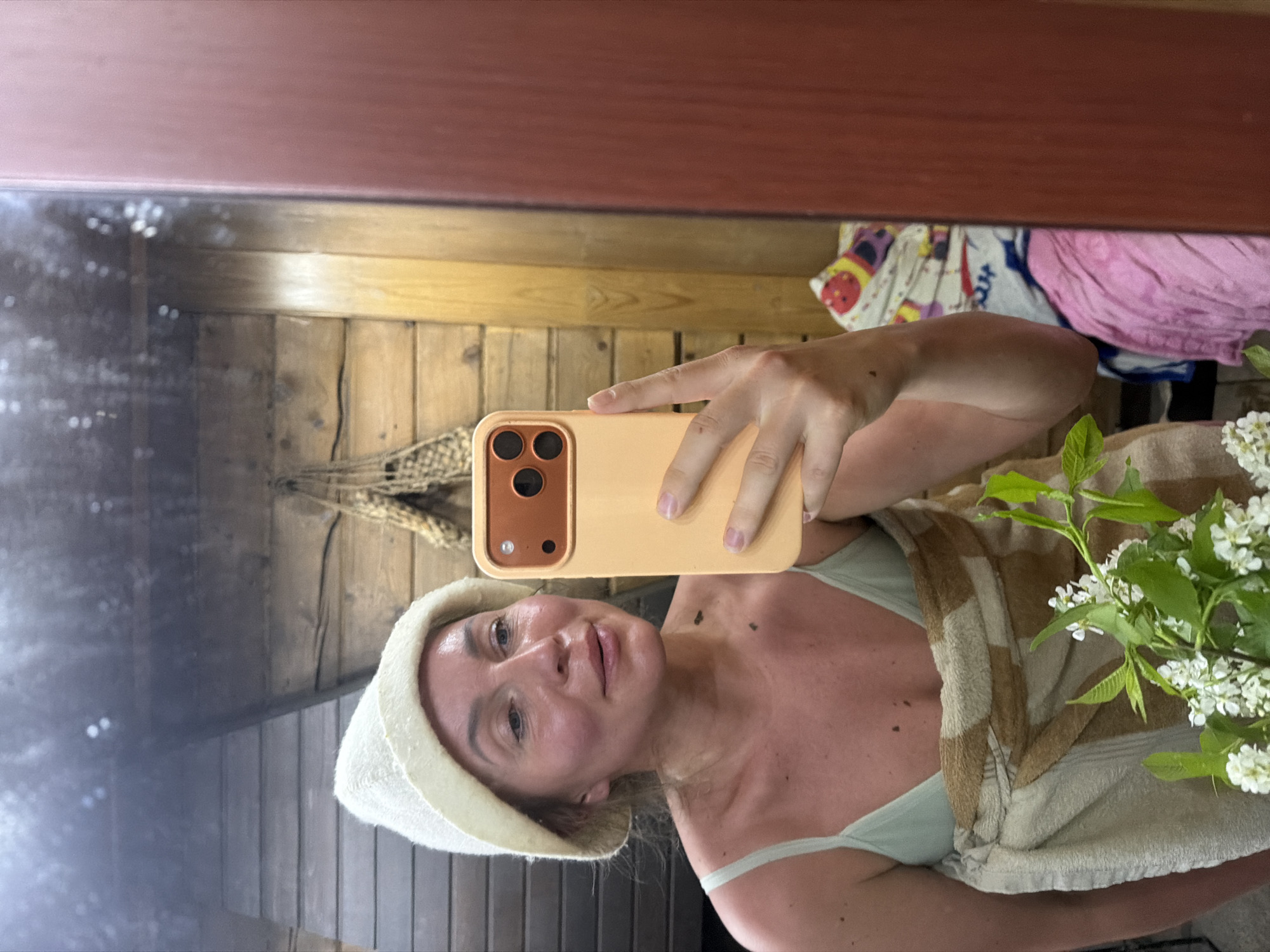 A woman is taking a selfie in what appears to be a sauna or bathhouse in Фоки, Russia. She is wearing a white felt sauna hat and a light green bathing suit top. A brown and white striped towel is draped around her. She is holding a light yellow smartphone, partially obscuring her face. In the background, there are wooden walls and a braided hanging decoration. A bouquet of white flowers is also visible. There are several other objects in the reflection.