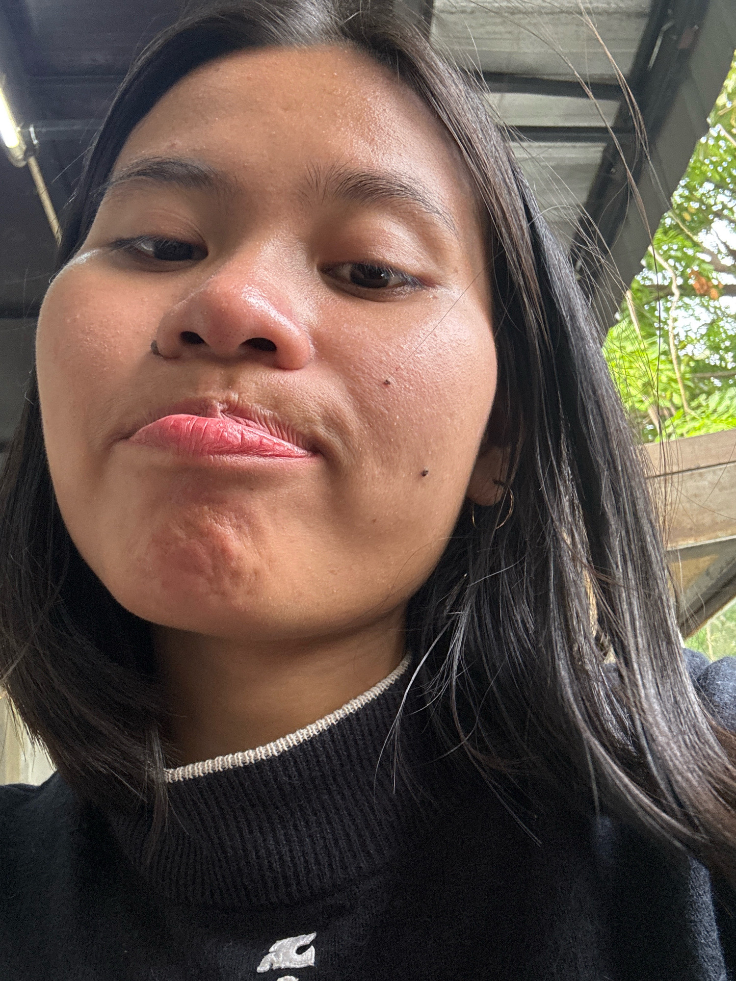 A medium shot shows a young woman's face filling most of the frame, angled downwards and slightly to her left, as if taking a selfie. She has dark hair that falls straight down around her shoulders and is wearing a black turtleneck sweater with a white trim at the neckline. Her expression is pensive, with her lips slightly pursed. The background appears to be outdoors, with hints of green foliage and a metallic roof structure visible above her head. The lighting suggests it might be daytime, possibly under an overhang. The location is East Jakarta, Indonesia.