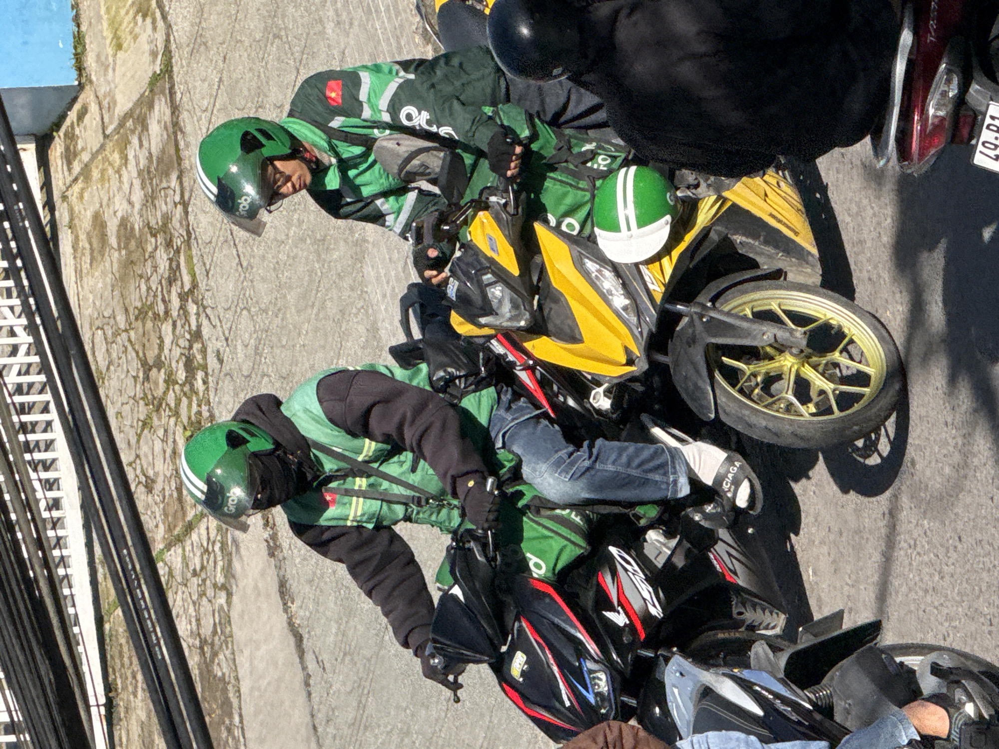 Two male individuals wearing green Grab delivery uniforms and helmets are on motorcycles on a paved road in Da Lat, Vietnam. The individual on the left is wearing a black face mask and is on a black and red motorcycle. The individual on the right is wearing glasses and is on a yellow motorcycle. Both individuals appear to be stopped. The environment includes a textured concrete ground with shadows cast by overhead structures and vegetation. There is a building with blue and white sections in the background.