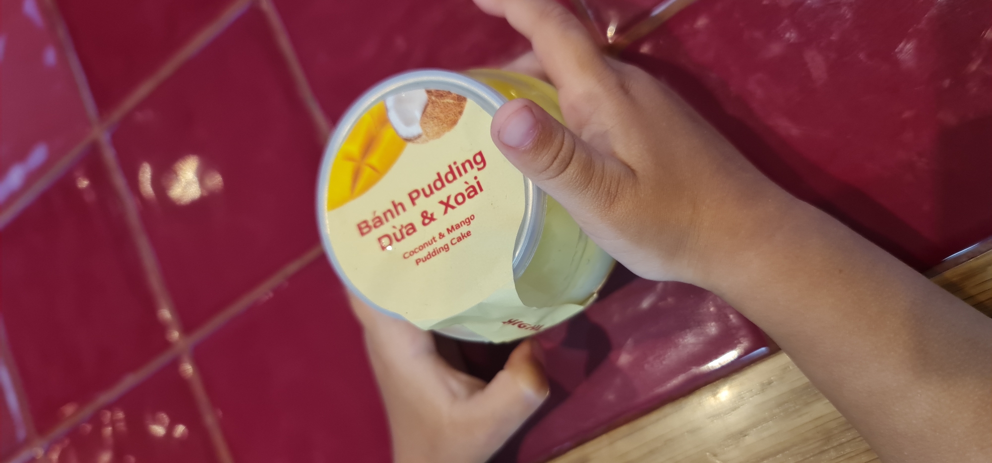 A child holds a container of Bánh Pudding Dừa & Xoài (Coconut & Mango Pudding Cake) in their hands. The container's label is predominantly yellow, with the pudding visible inside. The child's hands are light-skinned. The background consists of a red tiled surface and a wooden surface. The location is likely in Đà Nẵng, Vietnam, based on the product name.