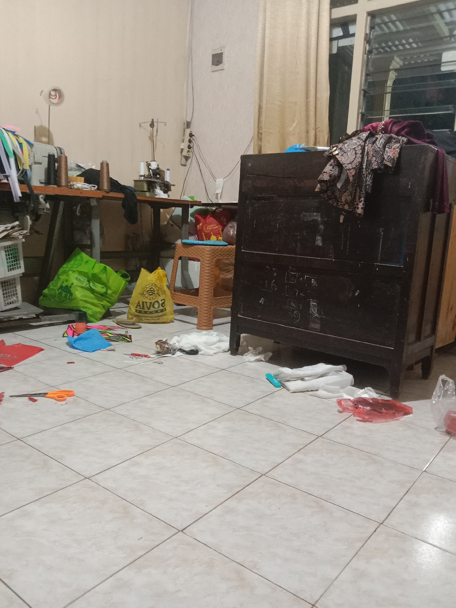 The image depicts a cluttered room, likely a workshop or storage area, with a tiled floor. A sewing machine is visible on a table in the background, along with spools of thread and various fabric scraps. Several plastic bags, both empty and containing items, are scattered across the floor, along with other discarded materials such as scissors and colorful paper. A dark wooden cabinet stands prominently in the foreground, adorned with a pile of patterned fabric at the top. The room appears to be dimly lit, with a beige curtain covering a window in the background. The overall impression is one of disarray and ongoing activity.