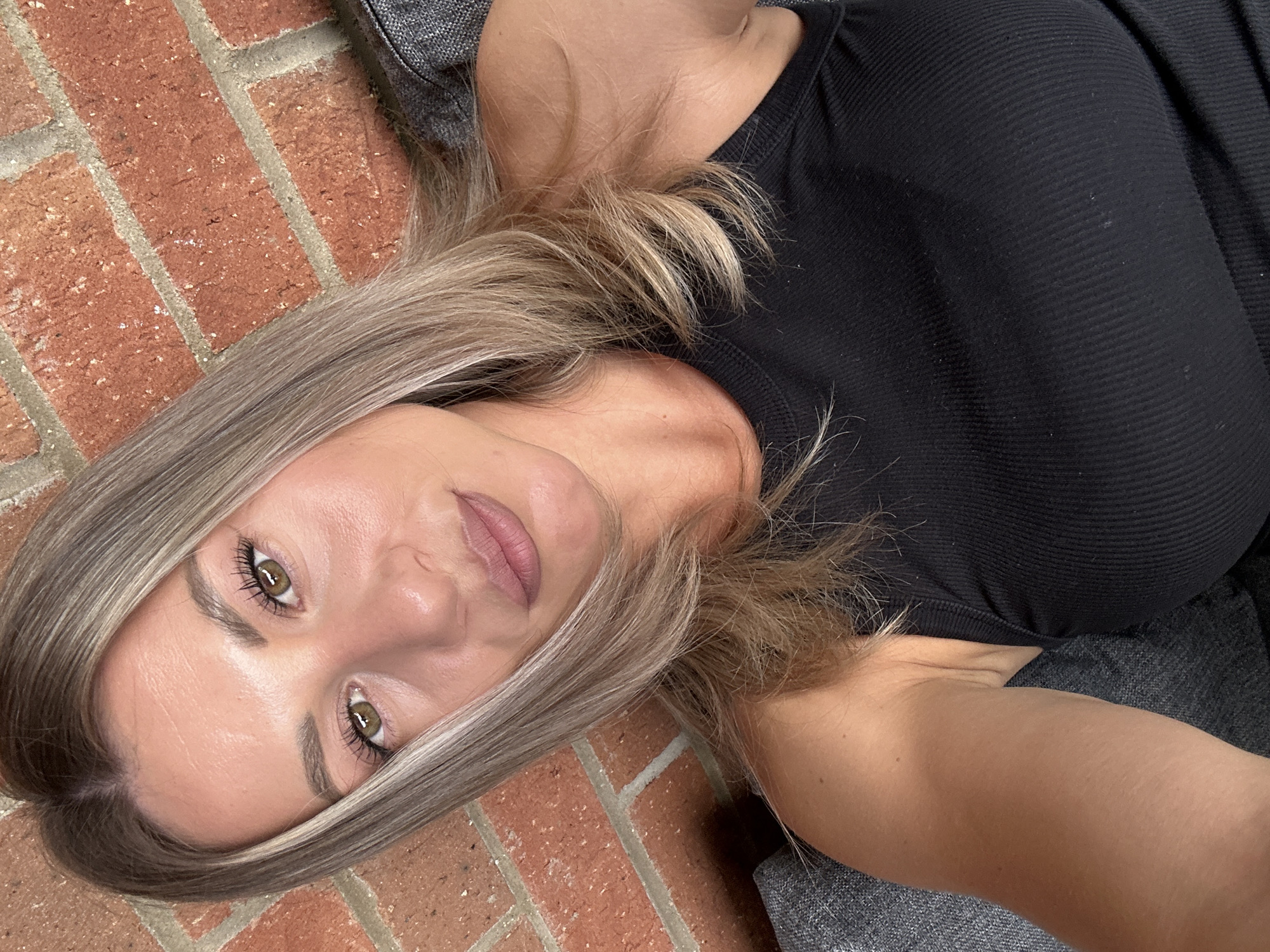 A woman with shoulder-length blonde hair, light brown eyes, and fair skin is pictured in a close-up selfie. She is wearing a black ribbed tank top and is seated on a grey sofa against a brick wall. The image shows her face, shoulders, and upper chest. The background consists of the textured red brick wall and the grey fabric of the sofa. The lighting appears to be natural, coming from above and to the side, casting subtle shadows on her face and neck. The overall impression is a casual, indoor portrait. It is not possible to determine the exact location in the city of Milton Keynes from the image.