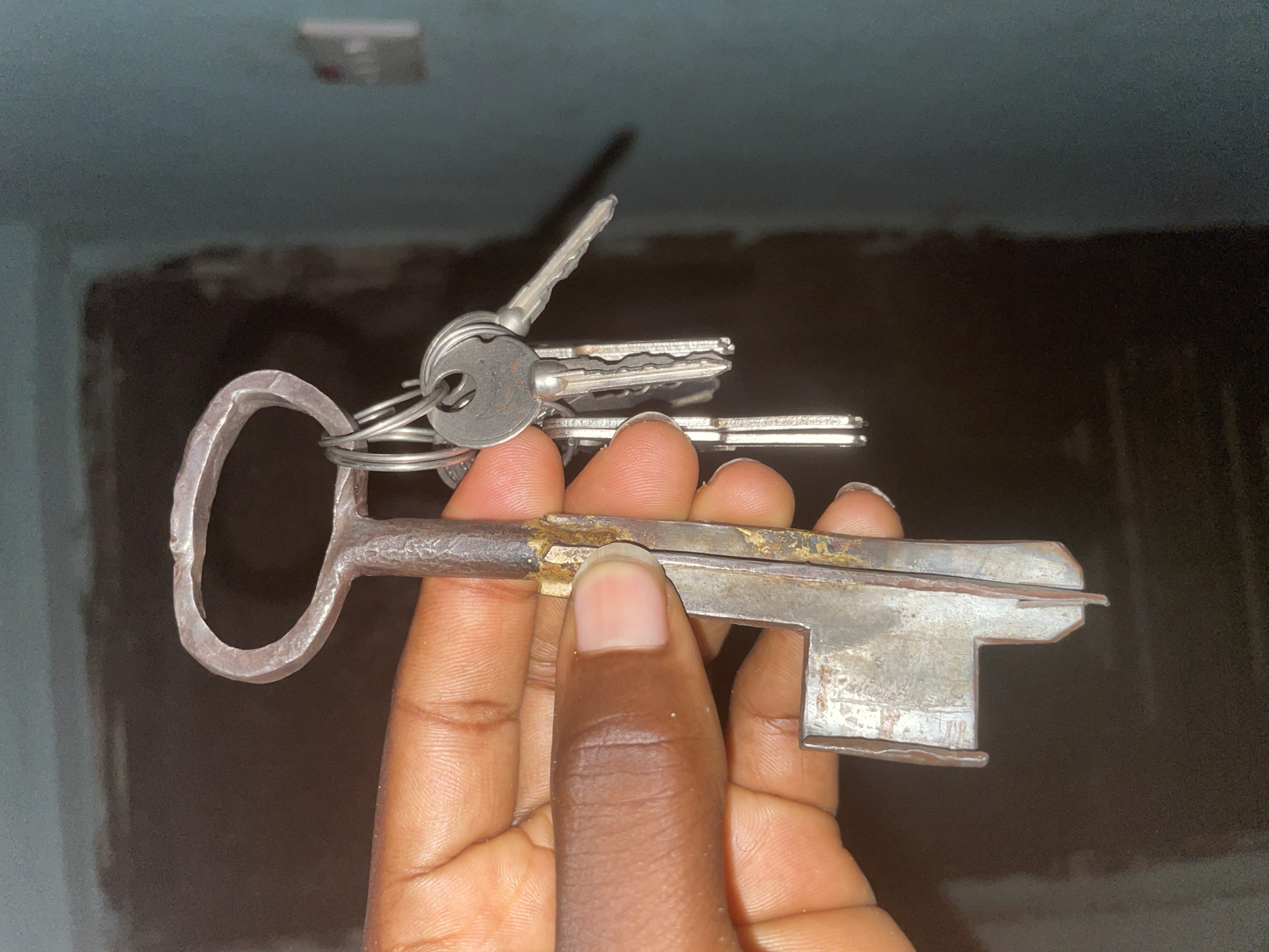 A person's hand holds a large, antique-style metal key with a loop at the top, from which hang several smaller, modern keys on a ring. The keys are made of a dull silver-colored metal, with the larger key showing some rust and discoloration. The person's skin tone is dark. The background is a dark, out-of-focus interior space with what appears to be a blue-painted wall on the right. This scene is set in the city of Jalingo, Nigeria.