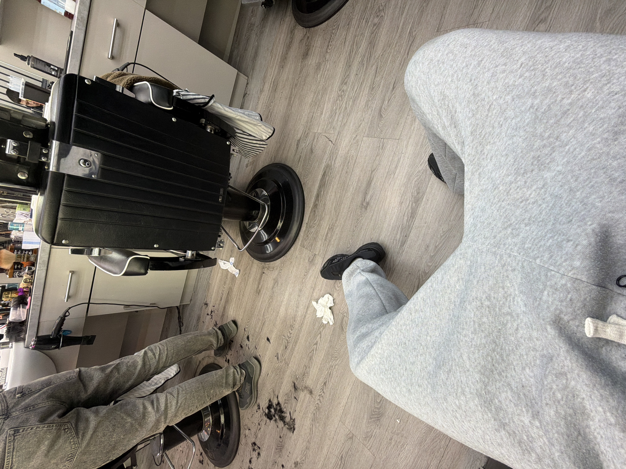 The image depicts an indoor scene, likely a barbershop. Two individuals are partially visible. One is wearing light grey sweatpants and black sneakers, their legs extending into the frame. The other person is sitting in a black barber chair, wearing grey jeans and green-grey sneakers. Hair clippings are scattered on the light wood-look floor. A black barber chair dominates the left side of the frame, with a white cabinet in the background holding various items, including hair products. Paper towels are discarded on the floor. No information suggests a location in London, United Kingdom.